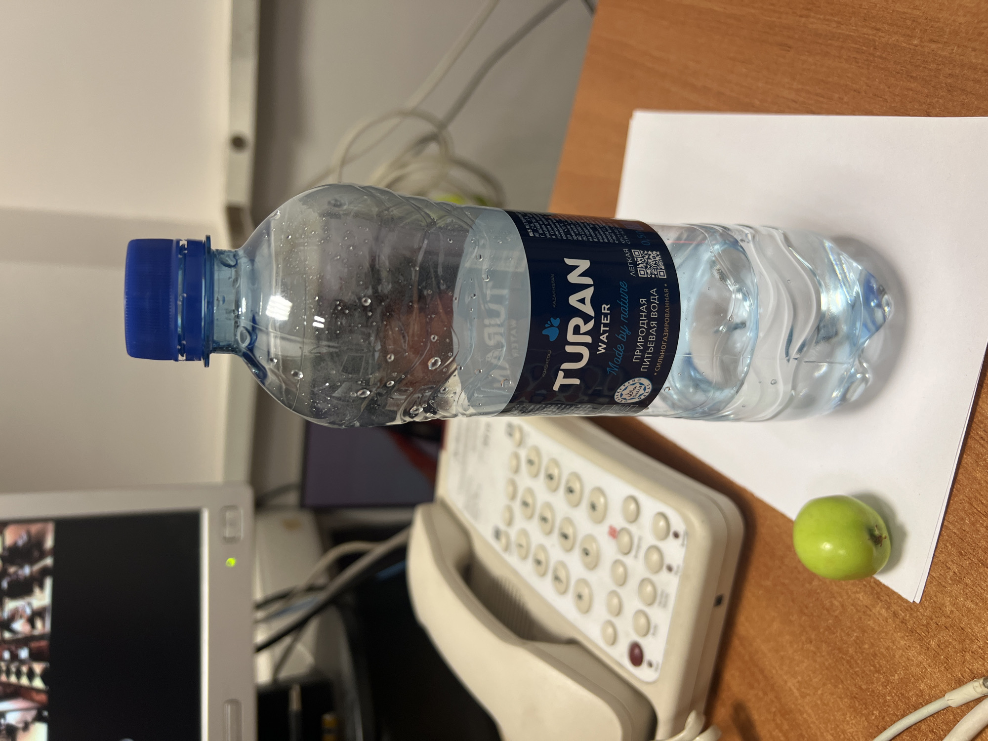 The image shows a desk scene in an office environment. The primary focus is a partially filled bottle of "TURAN WATER" with a blue cap, positioned on a white piece of paper. The label on the bottle indicates it is natural drinking water and made by nature, with text in Russian and a QR code. The bottle has condensation on its surface. To the left of the water bottle is a beige office telephone with a keypad and handset. In the foreground, near the bottom left, a small green apple sits on the desk. In the background, a computer monitor displays a blurred image, and various cables are visible. The desk surface appears to be made of wood. There is no indication of the specific location within the city of Astana, Kazakhstan, or any people in the image.