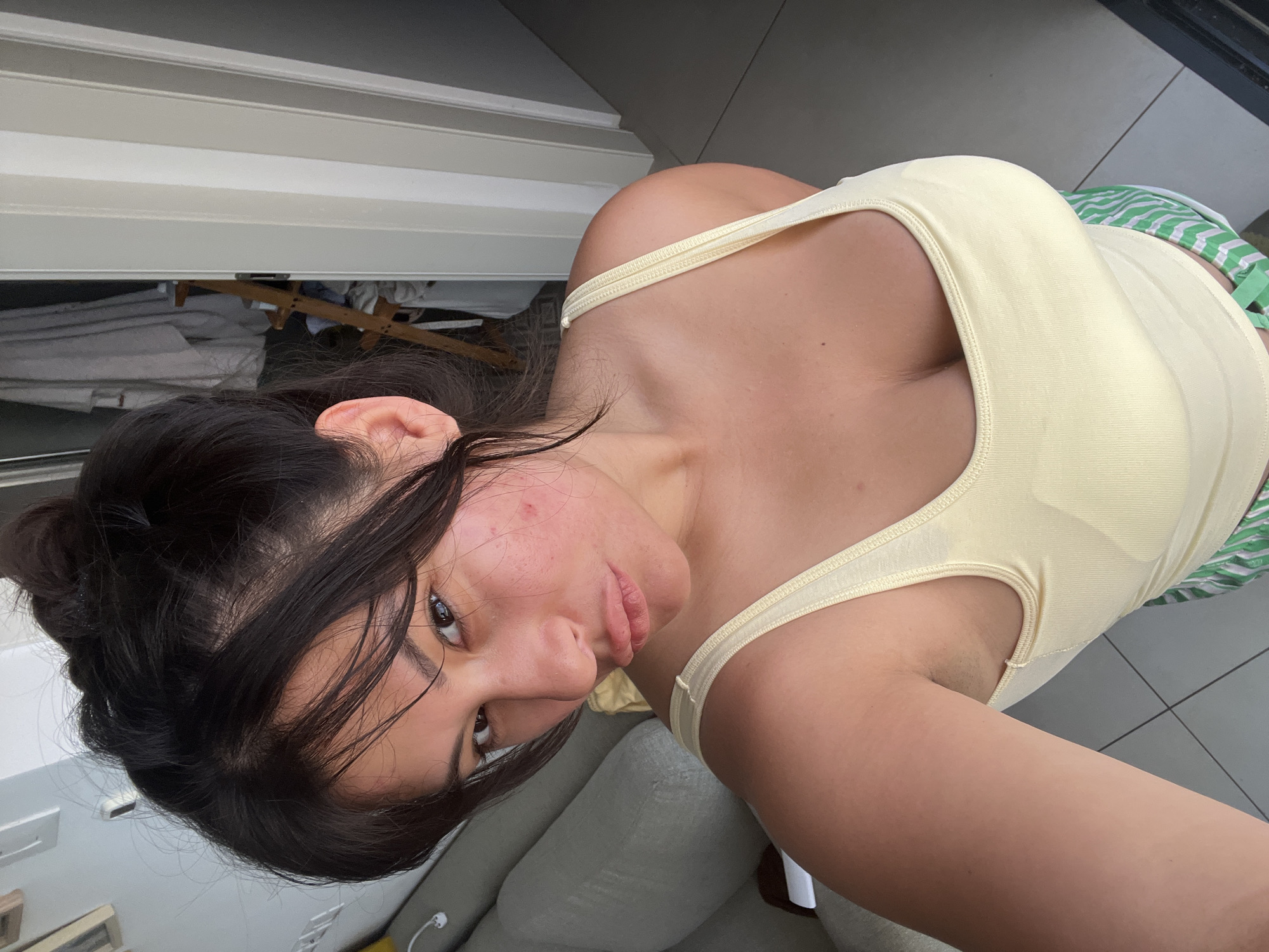 The image shows a young woman, likely in her late teens or early twenties, taking a selfie. She has dark hair pulled back in a bun, with some strands falling around her face. Her skin appears to have some redness and blemishes, particularly on her cheeks. She is wearing a light yellow tank top with thin straps and green and white striped shorts. The background is slightly out of focus, but it appears to be an indoor setting, possibly a living room or balcony, with tiled flooring and some furniture. There's a glimpse of a white door or wall with horizontal paneling on the left side of the frame. The lighting suggests it could be daytime. The woman's expression is somewhat serious, with her gaze directed at the camera. There is no visible text in the image. The location context provided is Kiryat Bialik, Israel.