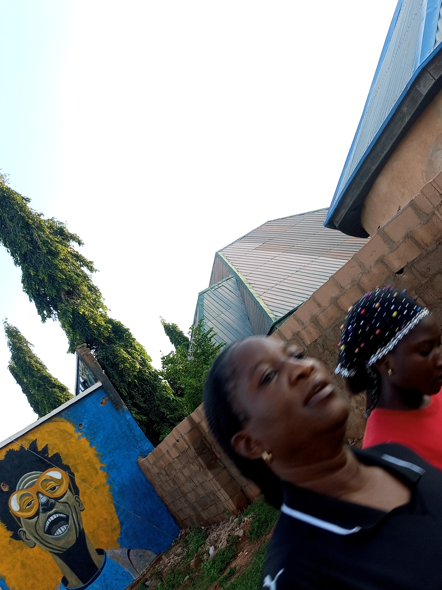 In Gombe, Nigeria, on a bright, possibly overcast day, two young women walk along a street. The woman in the foreground, with dark skin and hair pulled back, wears a black polo shirt with a white stripe. Her face is turned upward and slightly to the side, with her mouth open, as if speaking or reacting to something. She has small gold earrings. Behind her, and to the right, a second woman, also with dark skin and hair, wears a black headscarf with colorful polka dots and a red t-shirt. She is looking down and away from the camera, her facial features less distinct due to the blur.
The background reveals a vibrant mural painted on a wall, depicting a stylized portrait of a person with large, orange sunglasses and a bright yellow afro. The mural is set against a backdrop of blue. To the right of the mural and women, a brick wall is visible. Further back, tall, green, conical trees stand against a pale sky. In the upper right of the image, the slanted, corrugated metal roof of a building, painted blue along its edge, is prominent. The overall scene suggests an outdoor setting, perhaps a street or pathway in a residential or commercial area of Gombe. The angle of the shot is tilted, giving a dynamic perspective.