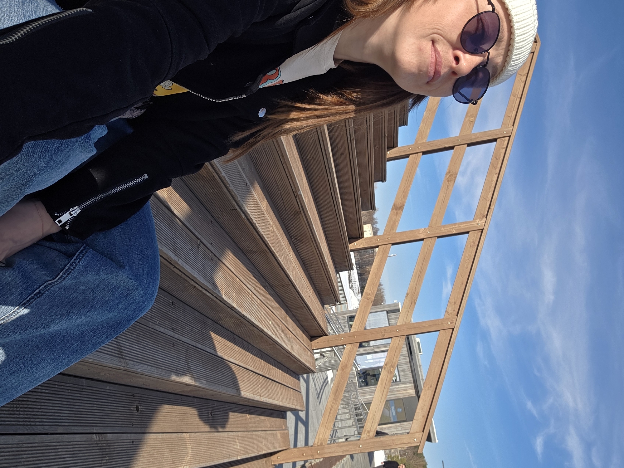 The image displays a female individual seated on wide wooden steps in Votkinsk, Russia, under a clear blue sky with sparse cirrus clouds. The subject occupies the lower-left quadrant, oriented slightly upward and to her left. She wears a white ribbed knit beanie, dark-tinted round sunglasses with black frames, a black zip-up jacket over a partially visible graphic t-shirt, and blue denim jeans. Her long, light brown hair cascades over her shoulders.
The steps are constructed from light brown composite wood planks and extend from the foreground into the mid-ground, forming a gradual ascent. A wooden railing with horizontal and diagonal slats runs parallel to the steps, separating the immediate foreground from the background. In the mid-ground, a contemporary building with a wooden facade and large glass windows is visible, featuring a sign with the Cyrillic word "ВЫХОД" (EXIT). To the right of this building, another individual in dark attire is partially visible walking on a paved surface. Behind the wooden railing, a silver metal handrail from an additional ramp or staircase is discernible. The background features distant leafless trees and a body of water, possibly Votkinsk Pond, with remnants of snow or ice on its banks. Strong shadows are cast across the wooden steps and railings, indicating direct sunlight.