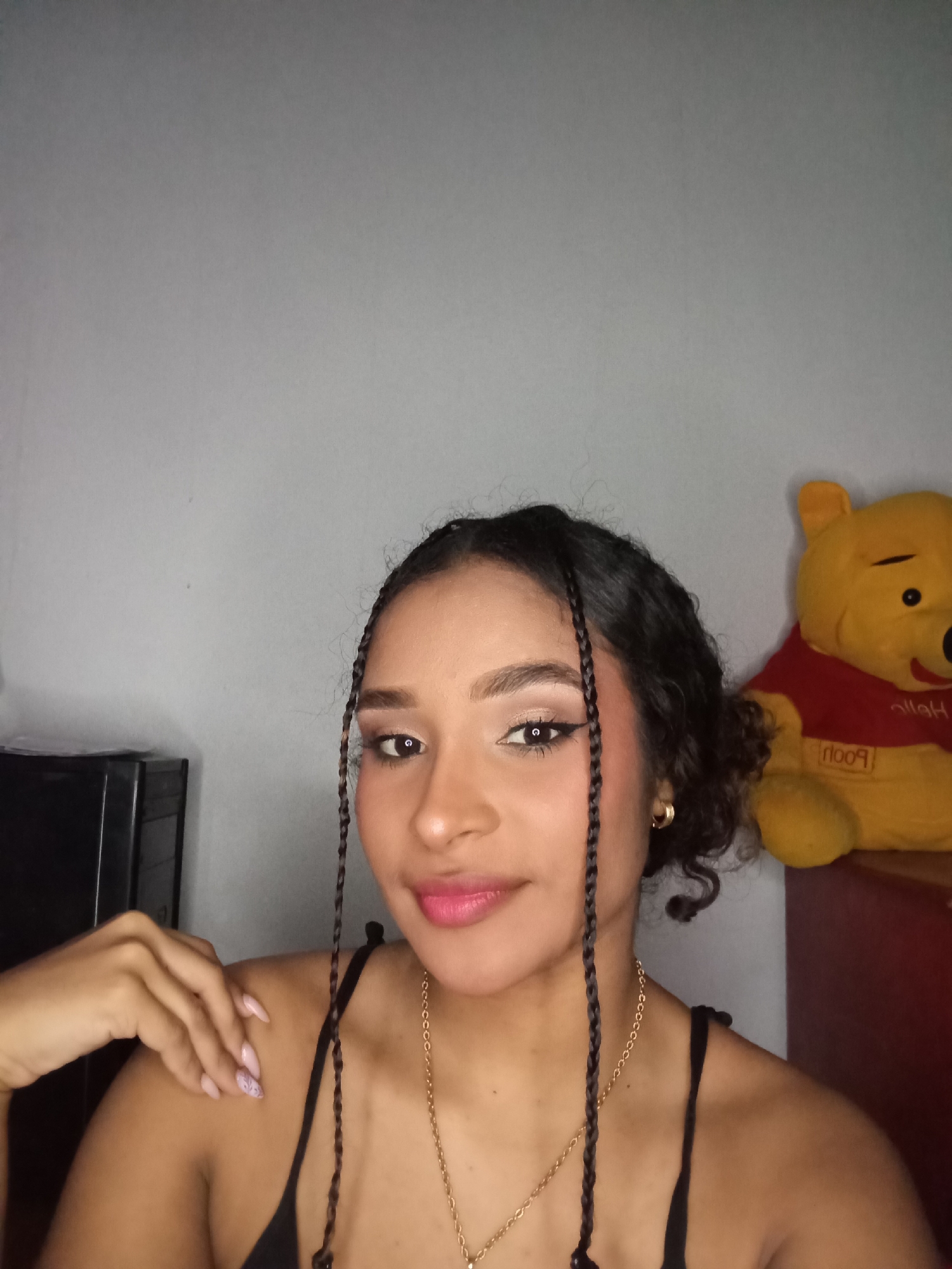 An indoor portrait image taken in Juan Griego, Venezuela, depicts a female subject positioned slightly off-center. The female has medium skin tone, dark, curly hair styled with several thin braids framing her face. She wears a black spaghetti strap top, a thin gold chain necklace with a small pendant, and a small gold hoop earring in her visible right ear. Her face features pink lipstick, defined eyebrows, and winged black eyeliner with light eyeshadow. Her right hand, adorned with light pink manicured nails, rests on her left shoulder. She looks directly towards the camera with a slight smile. The background consists of a plain, light grey wall. To the lower left, a portion of a dark, multi-drawer unit is visible. To the right, a yellow plush toy resembling Winnie the Pooh, wearing a red shirt with "Pooh" printed in reverse, is situated on a red surface. The scene is illuminated by artificial light.