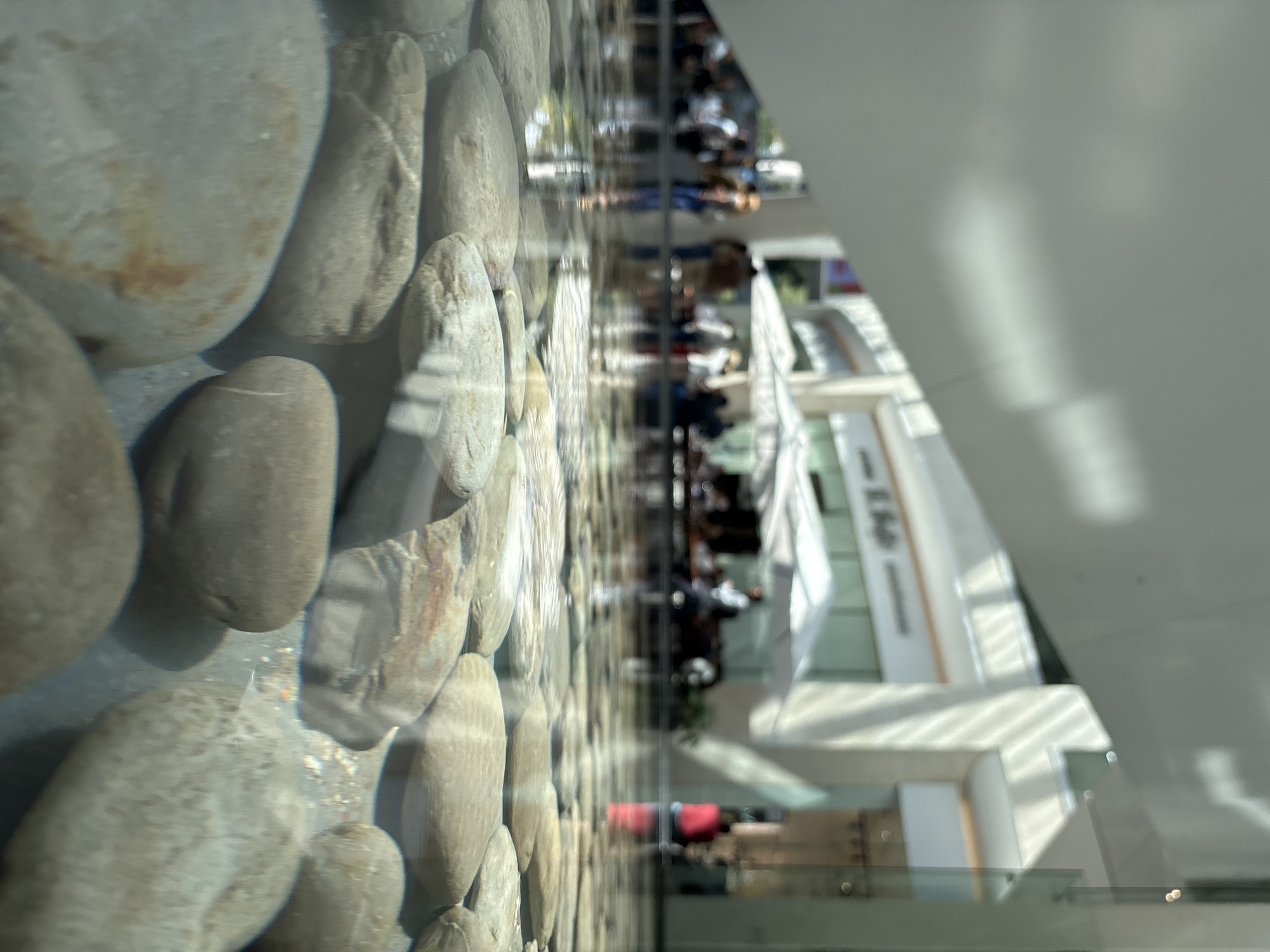 The image depicts a view through a clear glass partition or window, with a decorative element of rounded stones lining the inside of the glass. Through the glass, a bright, interior space is visible, possibly a shopping mall or atrium. There are people walking and sitting within this space, though they are blurred due to the focus on the stones in the foreground and the reflective nature of the glass. Architectural details visible include white walls, multi-level floors, and open spaces. The location is possibly in Mexico City, Mexico.