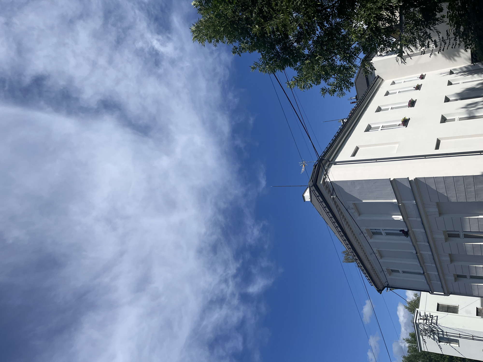 A low-angle, full shot captures a white, two-story building against a vivid blue sky with wispy white clouds. The building features multiple windows, some with small flower boxes. A dark, sloped roof tops the structure, with antennas visible. To the right, the dense green foliage of a tree partially obscures the building. Power lines stretch across the sky, and a smaller white building is visible in the background to the left. The scene appears to be an exterior shot on a clear day.