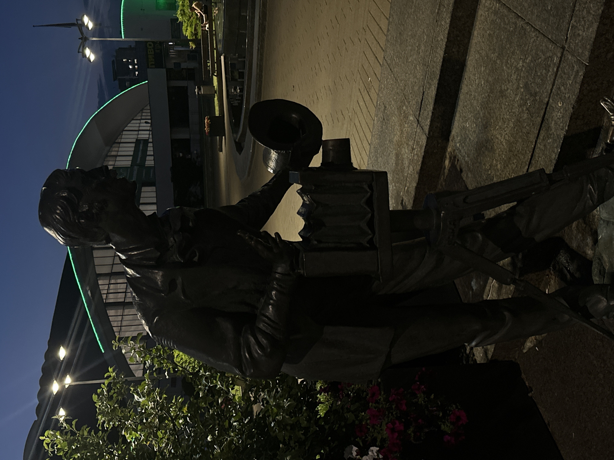 A full-body bronze statue of a man holding a vintage camera stands in a public outdoor space at dusk. The statue is positioned in the foreground, facing right, and is detailed with a suit, bowtie, and gloves. The camera appears to be a bellows camera mounted on a tripod. To the left of the statue is a planter box filled with green foliage and pink and white flowers. Behind the statue, a paved walkway with a brick-like pattern stretches into the background. Further back, a large, modern building with a curved, green-lit roof dominates the scene. Several streetlights with multiple bulbs illuminate the area. In the mid-ground, a public fountain is visible, and a few people are seated on a bench nearby. The sky is dark, indicating nighttime or late twilight. The overall impression is of a well-lit public square or park at evening.