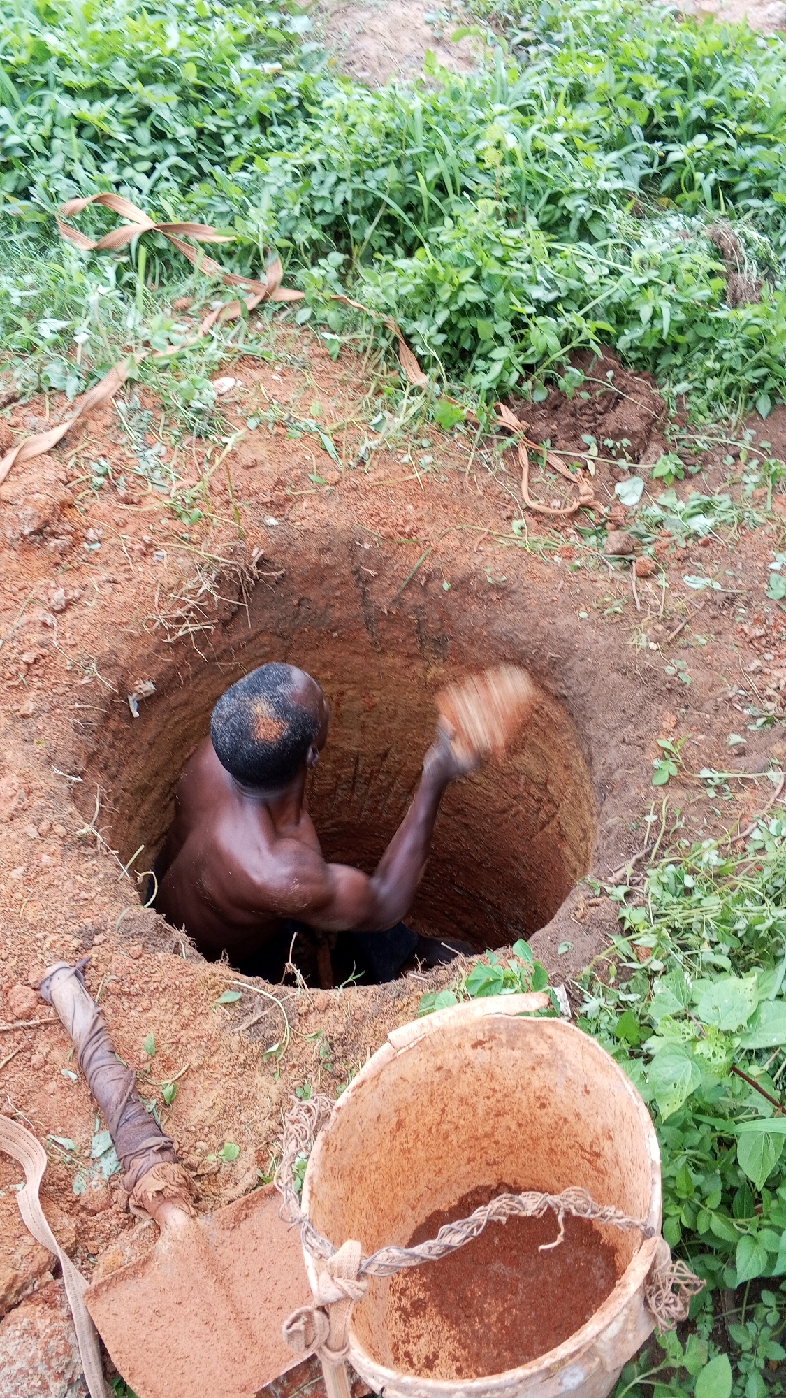 A man is digging a well in Bukuru, Nigeria. The man is inside the well, which is about 4 feet deep. He is using a shovel to dig the well. There is a bucket next to the well. The ground is reddish brown soil. The location is outdoors and surrounded by green vegetation.