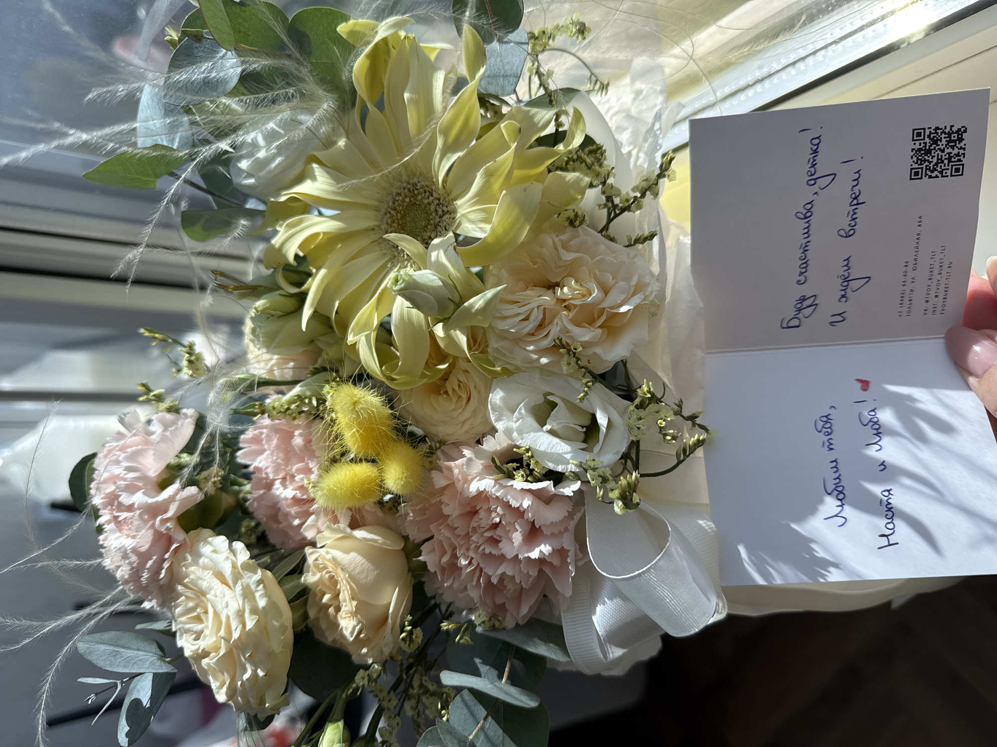 The image captures a bouquet of flowers. The bouquet consists of peach and cream-colored roses, carnations, eucalyptus leaves, and yellow straw-like flowers. The bouquet is accompanied by two cards. The first card is partially readable, and it contains the text: "Любим тебя, Настя и люба!". The second card is open and contains the text: "Буду счастлива, детка! И ждём встречи!". The second card also features a QR code and the address "ТОЛЬЯТТИ, УЛ. ЮБИЛЕЙНАЯ, 85A". The location is the city of Tolyatti, Russia.