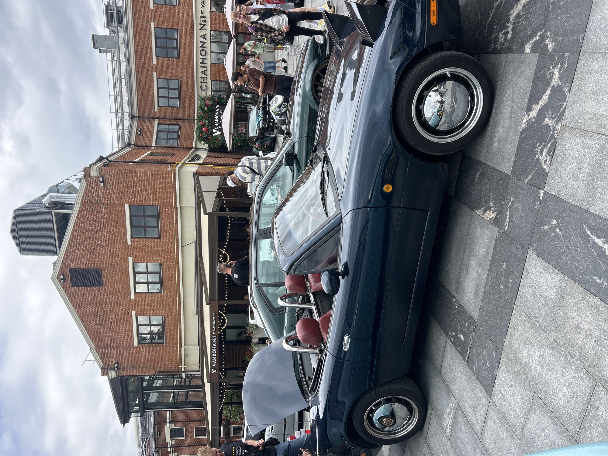 The image captures an outdoor scene in Moscow, Russia, likely at a car exhibition or gathering, given the presence of multiple cars and people. In the foreground, a dark blue convertible, possibly a Mazda Miata, is parked, its hood open, revealing the interior. The interior features red leather seats and a chrome roll bar. The car is on a paved area with large, grey stone tiles.
Behind the convertible, a dark green sedan is parked. Several individuals are seen milling around the vehicles and the surrounding area. One person in the foreground is leaning against a silver car, partially visible. Further back, a group of people are gathered, some looking at the cars.
The backdrop features a multi-story building constructed of red brick, with numerous windows. Above the building, a cloudy sky is visible. Signs on the building indicate businesses, including "CHAIKHONA №1" and "CHAIKHONA N°1 BY VASILCHUKOV", suggesting a restaurant or cafe is located there. The overall atmosphere appears to be casual, with people enjoying the display of vehicles.
