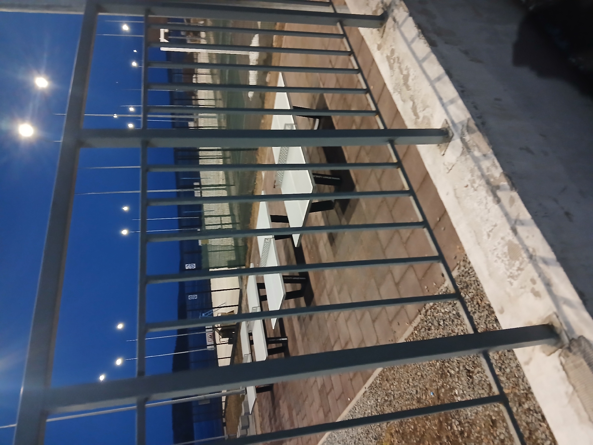 The image depicts an outdoor area in Chaikovsky, Russia, featuring metal railings in the foreground. Through the railings, a series of white picnic tables with black benches are visible, positioned on a surface comprised of paving stones and gravel. In the background, the sky is a deep blue, illuminated by a series of streetlights. The presence of the metal railings suggests a viewing platform or enclosed area overlooking the tables. The time of day appears to be evening or twilight, as indicated by the deep blue sky and the illuminated streetlights.