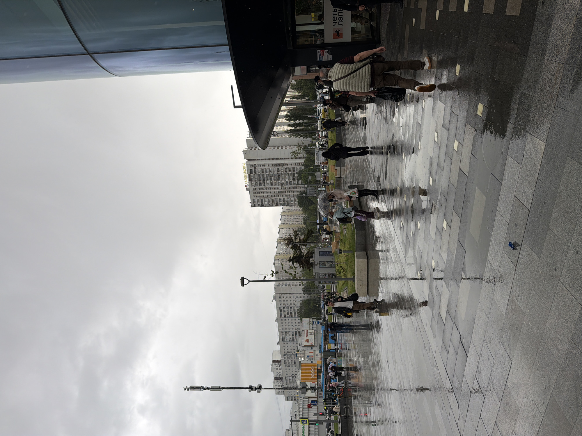 The image depicts a wet urban scene in Moscow, Russia. The camera is angled upwards, showing a view of a street with pedestrian traffic on a rainy day. People are walking on a paved area that reflects the surrounding environment due to the rain. Some individuals are carrying umbrellas. In the background, there are high-rise apartment buildings, indicative of a residential area in Moscow. Streetlights and signage are visible along the street. The sky is overcast. The image captures a typical rainy day in a city.