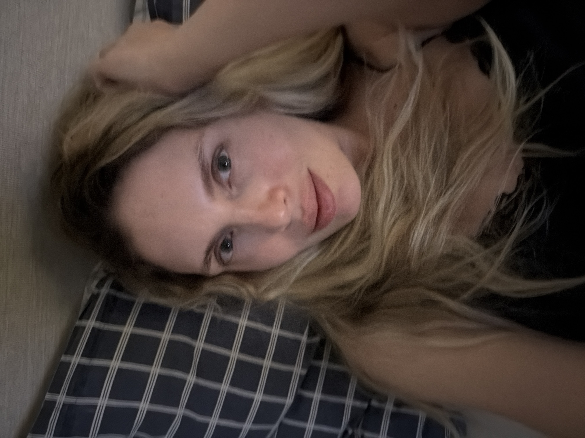 A woman with fair skin and blonde hair is lying on a pillow with a dark blue and white grid pattern. She is wearing a black top. The environment is not discernible enough to determine the location in Budva, Montenegro.