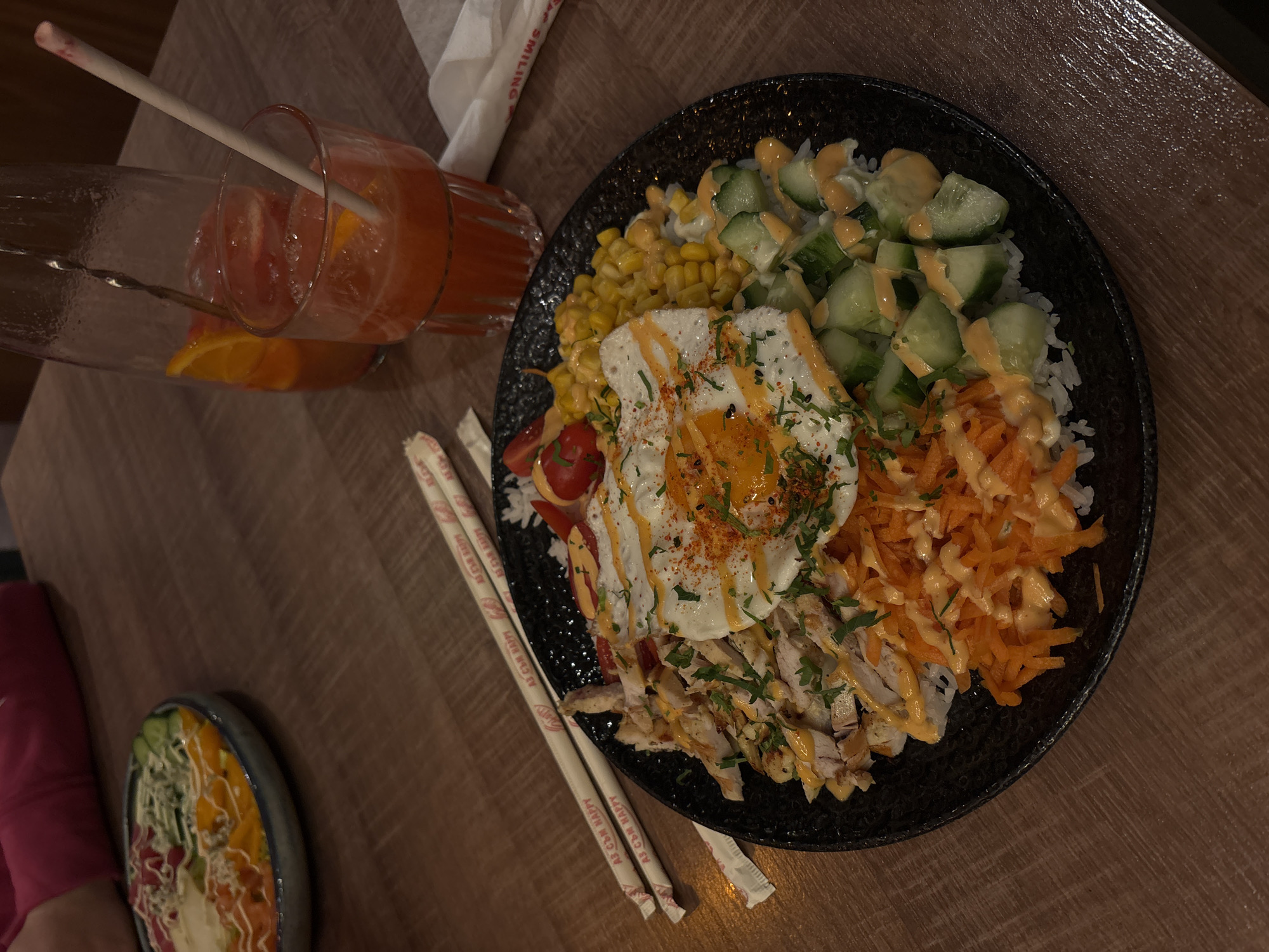 The image depicts a table scene, likely indoors, with a wooden surface. The primary subject is a black bowl filled with a diverse meal, featuring rice as a base, topped with corn kernels, diced cucumbers with sauce, shredded carrots, diced chicken, cherry tomatoes, and a fried egg with sprinkled paprika. Chopsticks are placed near the bowl, on the left. Beside the bowl is a tall glass with an orange-colored beverage, containing orange slices and a straw. A smaller bowl with a salad is partially visible on the left. The setting suggests a restaurant or dining establishment, possibly in Varna, Bulgaria. No people are fully visible, but a person's sleeve can be seen in the bottom left of the image.