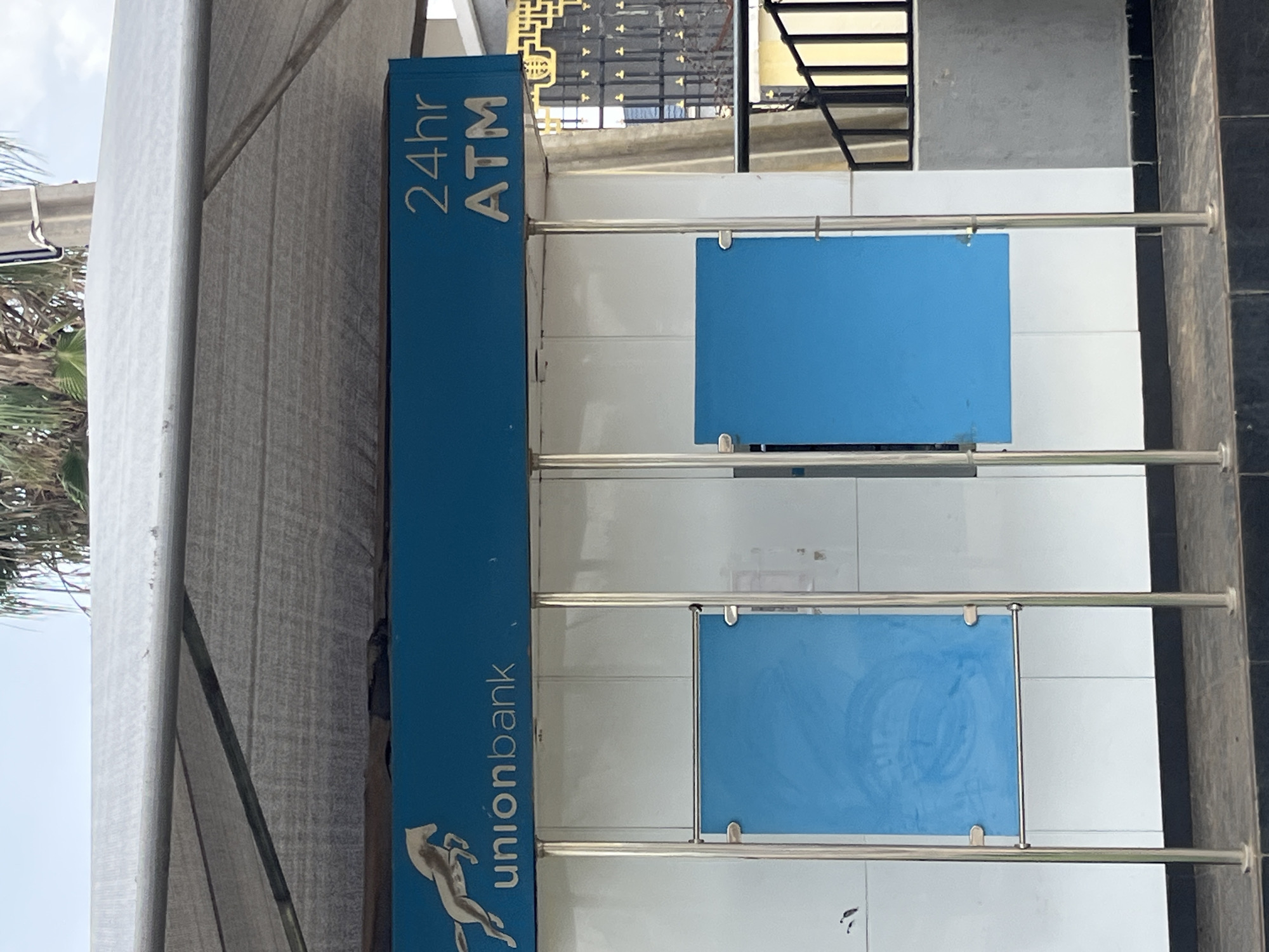 The image shows an outdoor ATM kiosk for Union Bank, located in Bukuru, Nigeria. The kiosk features a blue awning with the Union Bank logo, which includes a leaping horse, and "24hr ATM" text. Two blue panels are mounted on metal poles in front of the ATM machine, which is not visible. The kiosk is situated on a paved surface with dark gray and black tiles. In the background, a metal fence with decorative elements and a glimpse of a building with a patterned facade are visible. Palm trees and a bright sky suggest an outdoor, possibly sunny, environment. The overall scene depicts a standard banking service point in an urban or semi-urban setting.