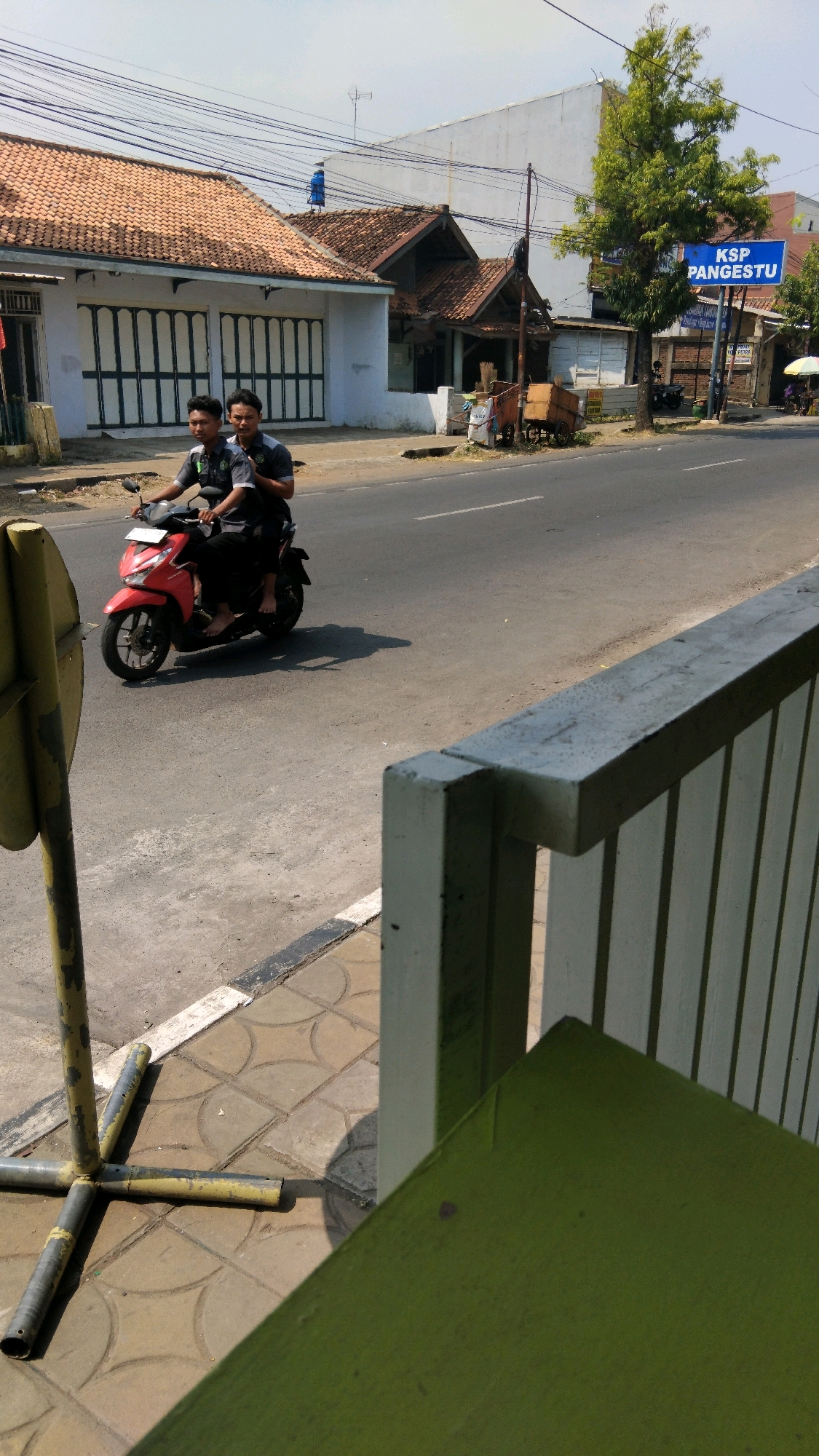 Two young Indonesian males are riding a red scooter on a paved road in Proyonangg, Indonesia. The rider in front wears a grey shirt with a logo, and the passenger behind him wears a similar shirt. Both are looking forward. The scooter is in motion, casting a shadow on the road. The scene is framed by a yellow signpost on the left, a white fence with vertical slats in the foreground, and buildings with tiled roofs on the right side of the road. A blue sign with white text reading "KSP PANGESTU" is visible on the right. The sky is overcast.