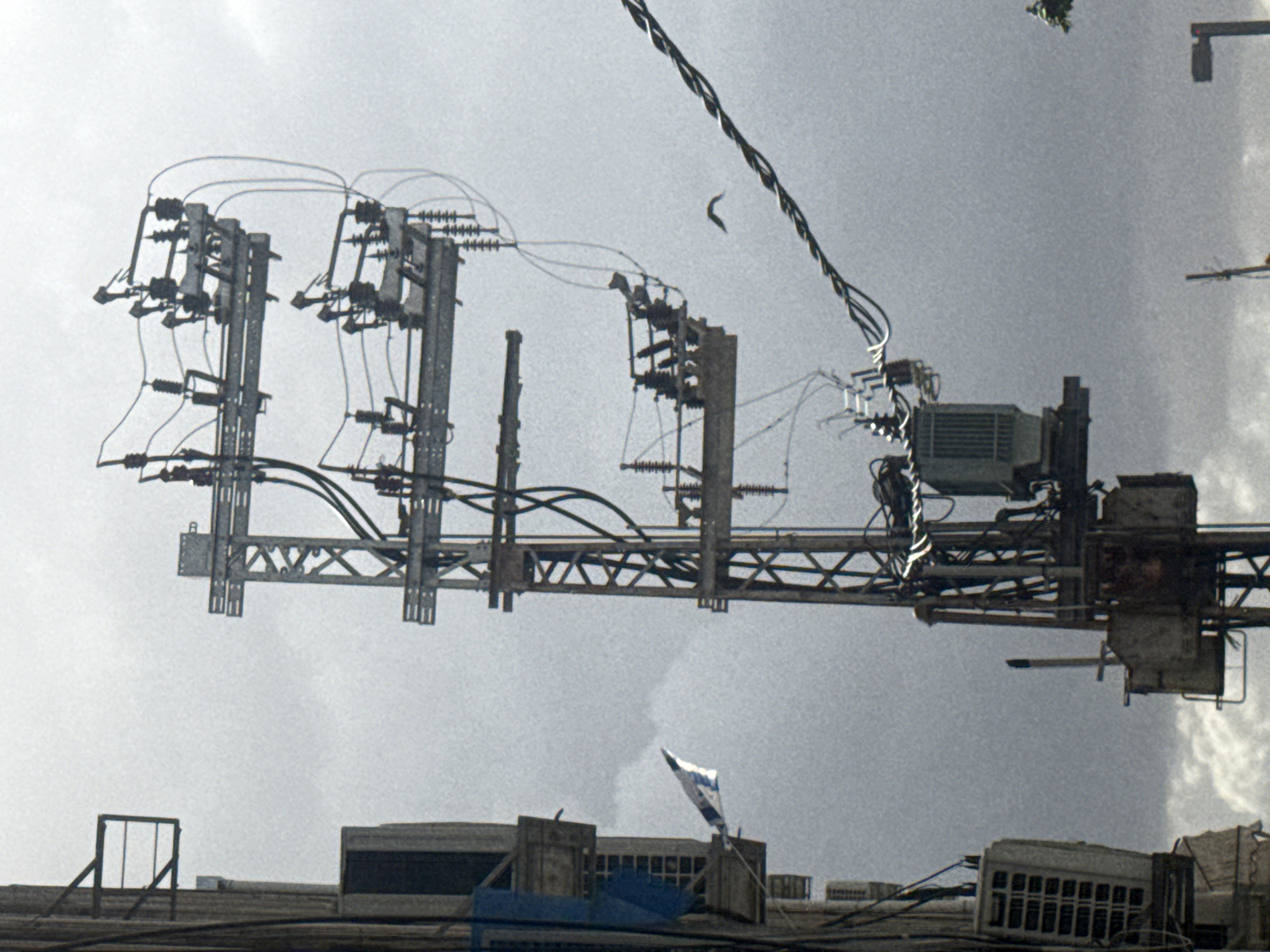 The image depicts a scene in Tel-Aviv, Israel, focusing on an infrastructure of electrical equipment suspended above a rooftop. Visible against a cloudy sky, the scene is dominated by metal structures carrying transformers, insulators, and power lines. The intricate network of wires and metal supports stretches across the upper part of the frame. Below the infrastructure, a building rooftop is visible, marked by various rooftop units and a waving Israeli flag. The overall scene appears utilitarian, highlighting the industrial aspect of the urban landscape.