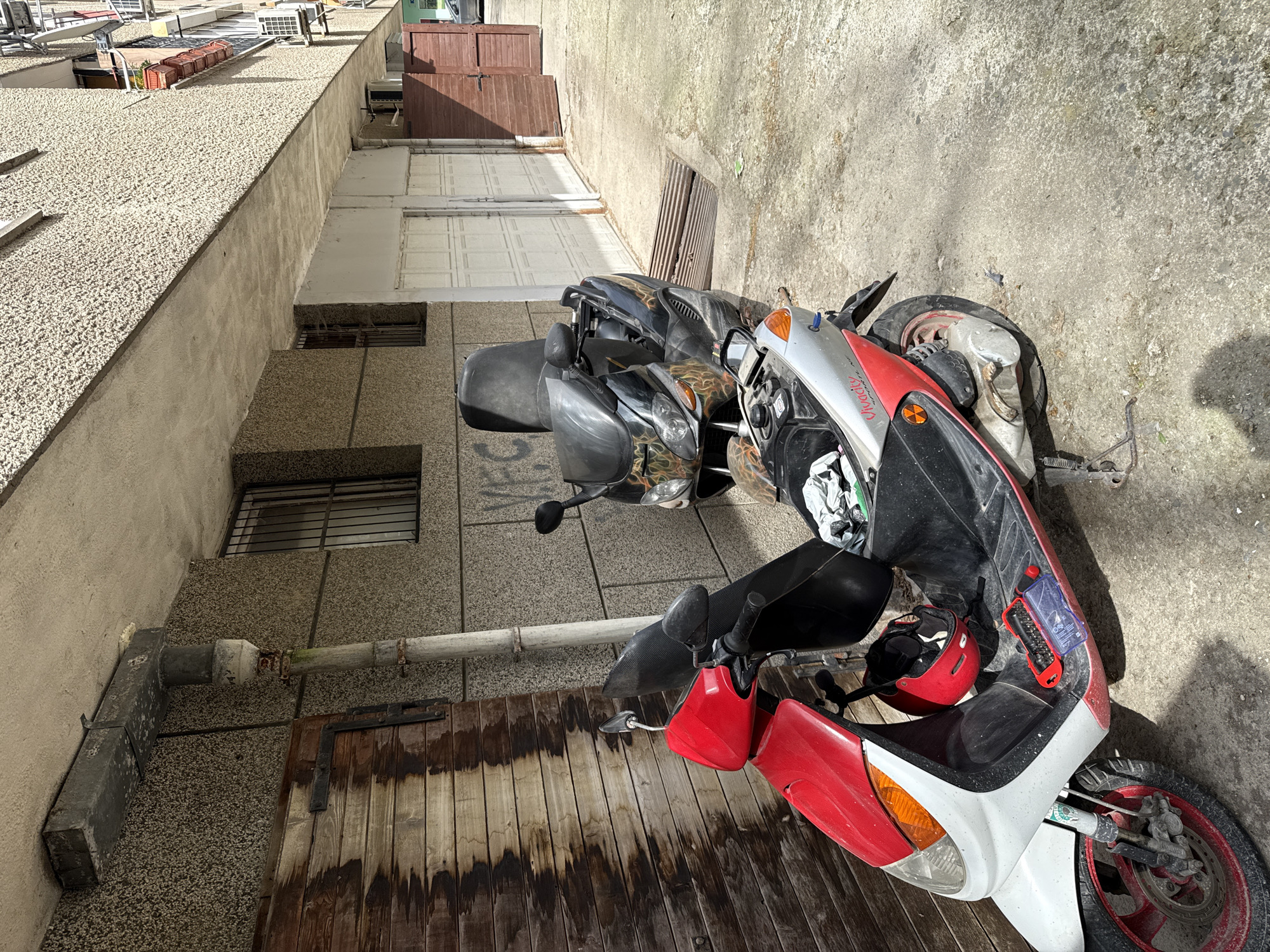 The image shows two parked scooters in an alleyway between buildings in Stara Zagora, Bulgaria. The alley is narrow, with a textured concrete wall on one side and a taller building with windows and doors on the other. The scooters are parked on a wooden surface, possibly a gate or platform. The scooter closest to the viewer is predominantly red and white, with black accents. The other scooter is black with a camouflage pattern. A red helmet is on the seat of the red and white scooter. The lighting suggests a sunny day. The surroundings include an urban environment with rooftops and building details visible above.