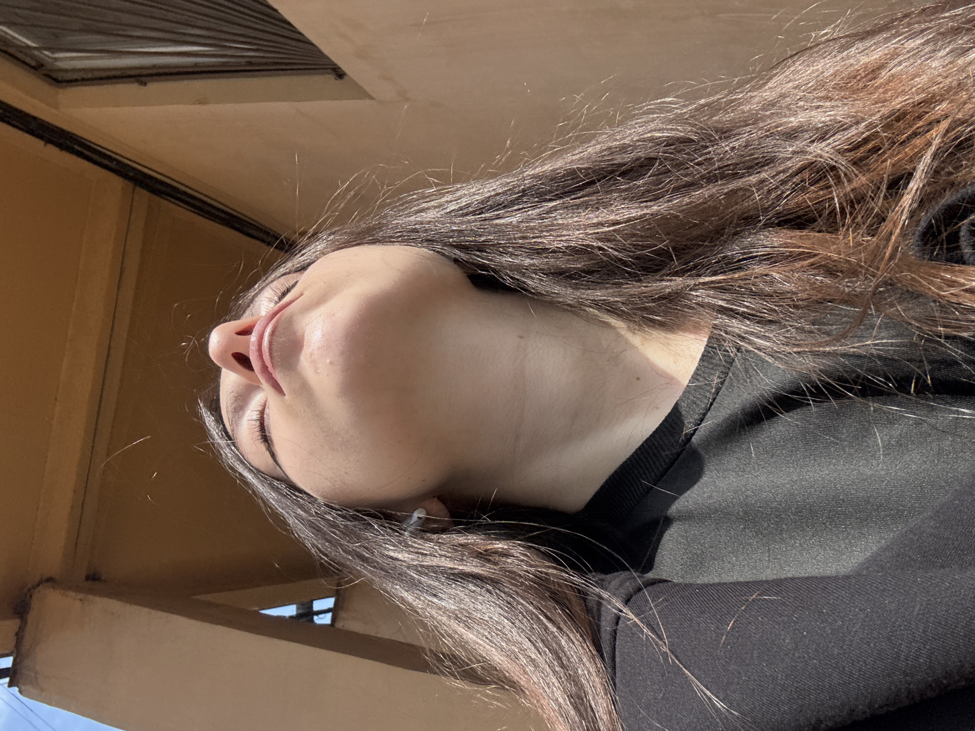 A low-angle shot depicts a woman with long, dark brown hair and closed eyes, lying beneath a beige architectural structure. The view captures her face, showing the nose, mouth, and closed eyelids. She is wearing a black top. The architectural structure appears to be a building overhang or balcony with beams. The location is not discernible from the visual information.