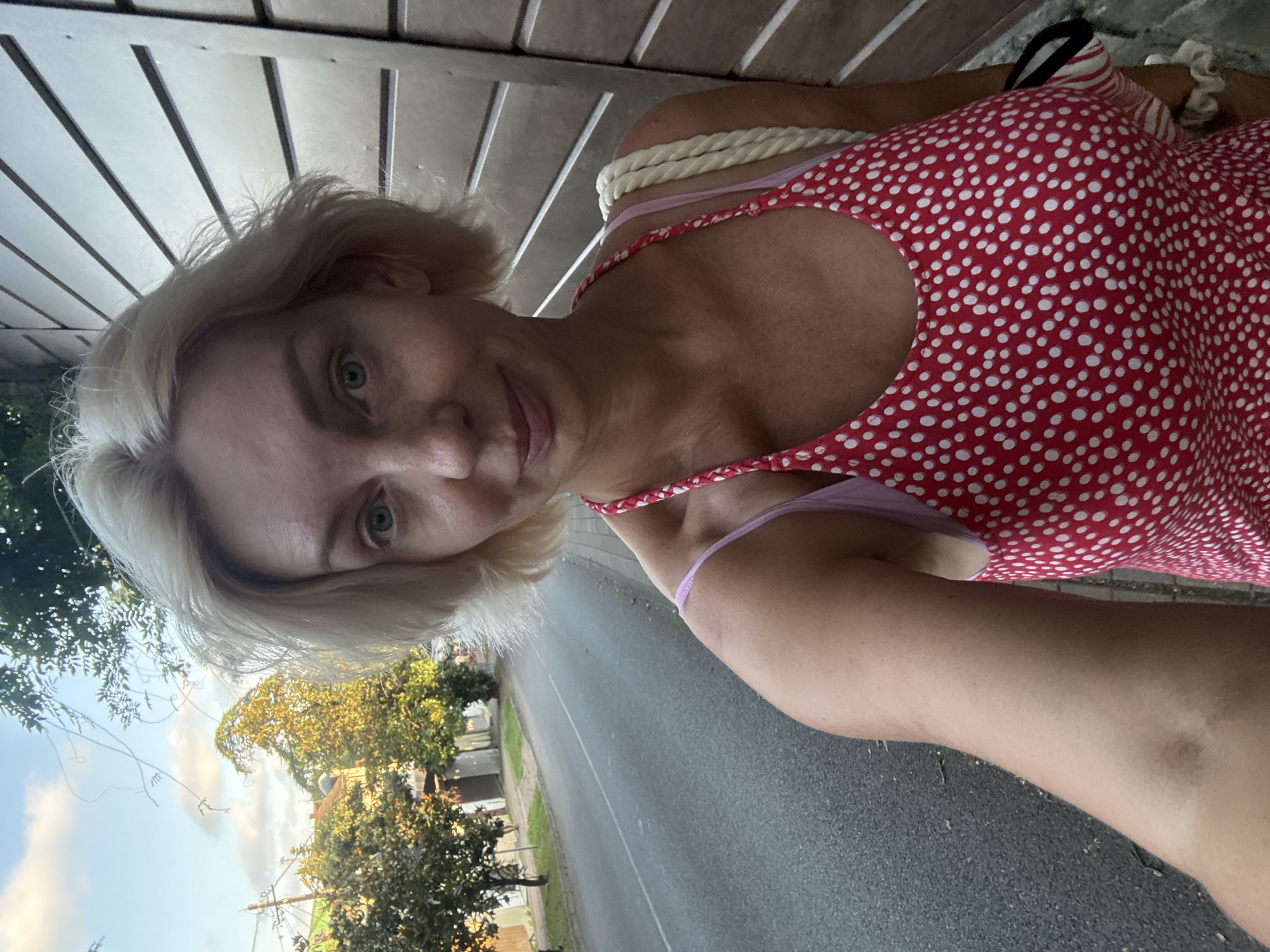 A woman with blonde hair stands on a sidewalk next to a road, looking at the camera. She is wearing a red dress with white polka dots and a pink bra underneath. She is carrying a white rope bag over her shoulder. Behind her is a wooden fence. The road curves to the left, with trees and buildings lining the other side. The sky is blue with some clouds. The image appears to have been taken in Sochi, Russia.