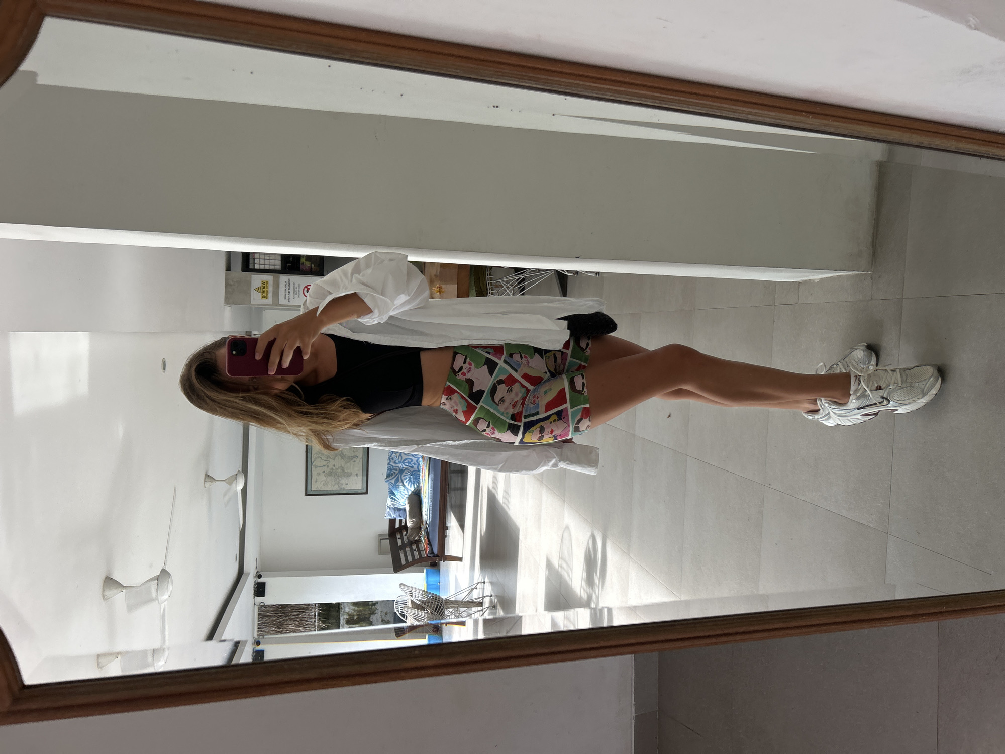 A female individual is captured in a mirror selfie in a well-lit interior space. She is dressed in a black crop top, a patterned skirt, and a white oversized shirt. The person holds a smartphone with a red case, obscuring her face. The reflection shows a part of a room with a table, chairs, and other furniture. Outside a window, there is greenery. The location is identified as Pererenan, Indonesia. The time of day appears to be during daylight hours.