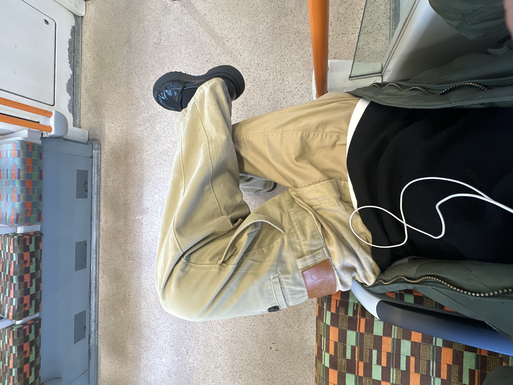 The image shows a person sitting on a seat in what appears to be a train carriage, likely within the city of London, United Kingdom. The person is wearing beige trousers, a black sweater, and a green jacket. They have black shoes on. The train carriage has a grey wall, orange handrail, and patterned seats. A white cable is visible near the person's chest.