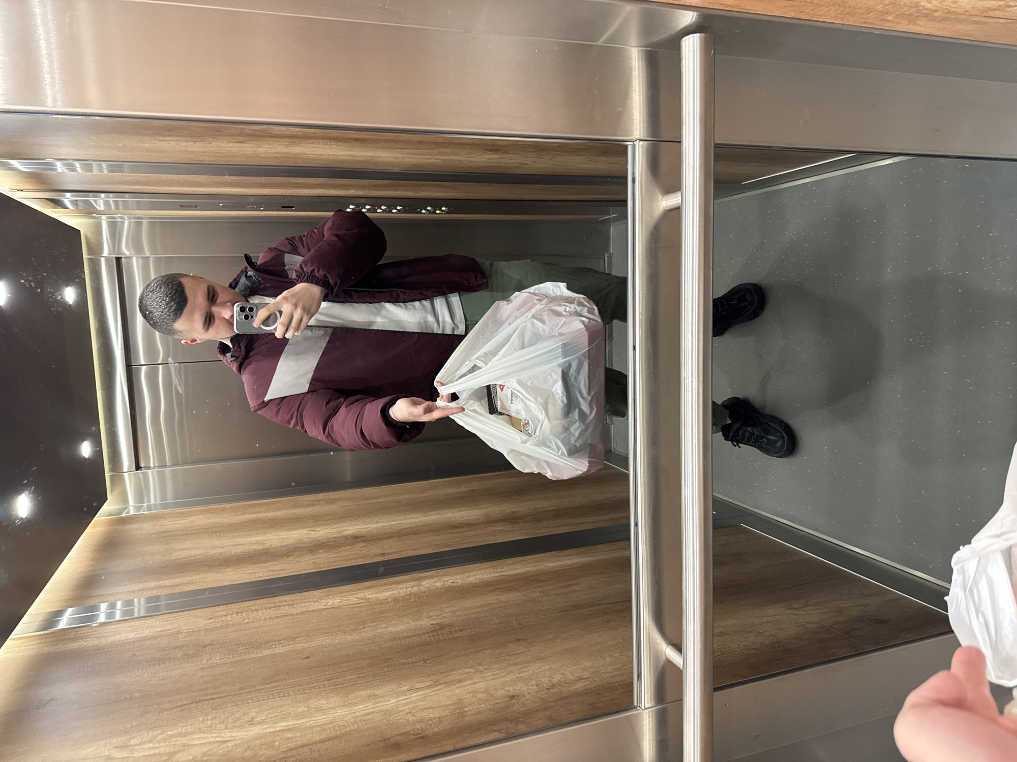 A man is standing in an elevator, taking a selfie in the mirror with his smartphone. He is wearing a maroon jacket, a white shirt, and green pants. He is holding a white plastic grocery bag. The elevator has wood paneling on one wall and a stainless steel and black panel on the other. The floor is gray. The image does not contain sufficient information to determine the specific location in Sofia, Bulgaria.