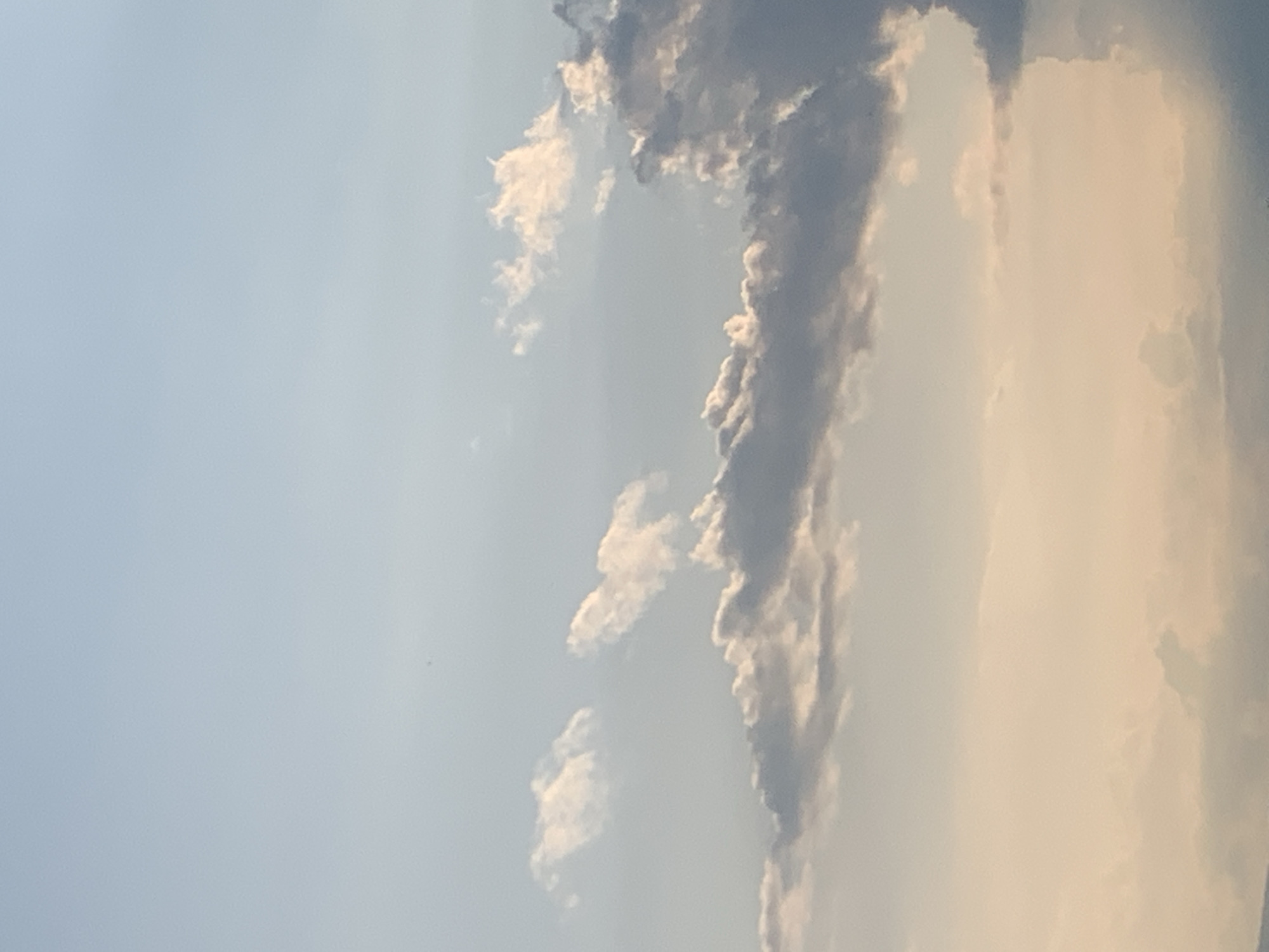 The image is a vertical, low-angle shot of a sky filled with clouds at various altitudes and densities. The sky transitions from a light blue at the top to a pale, warm orange-pink hue towards the bottom, suggesting either sunrise or sunset. Several distinct cloud formations are visible. In the upper half, small, wispy white clouds are scattered. A more prominent, elongated cloud bank runs across the mid-section of the image, with its upper edges illuminated and appearing white, while the undersides are tinged with gray and darker tones. Closer to the bottom, larger, more textured clouds are visible, some with darker gray undersides and brightly lit upper surfaces. There are no people, man-made structures, or recognizable landmarks visible in the image. The scene appears to be an open sky, devoid of any specific urban context, and thus cannot be definitively located within Kolomyia, Ukraine, or any other city.