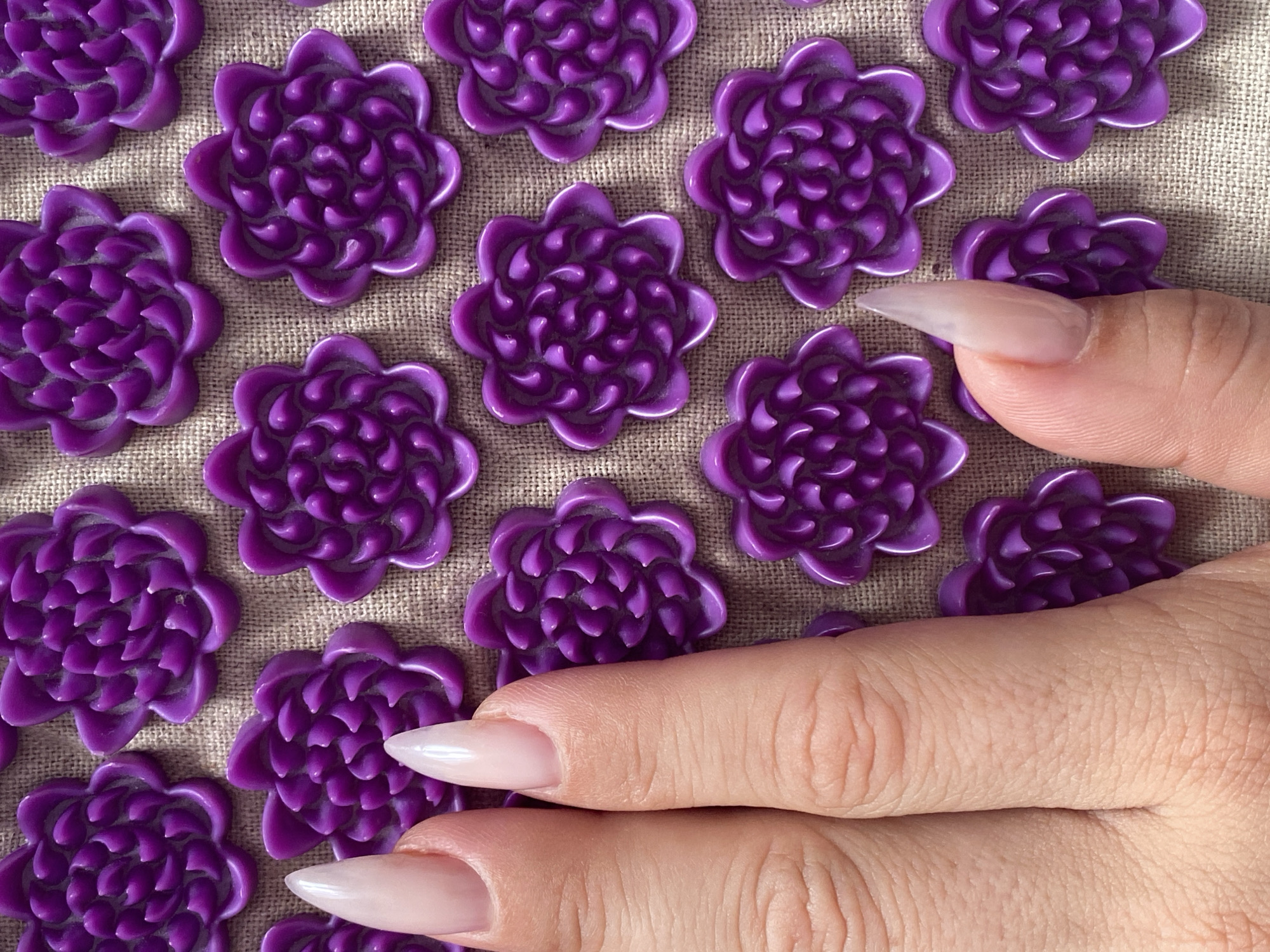 A close-up overhead shot shows a hand with long, almond-shaped acrylic nails resting on a mat. The mat is covered in a grid of small, purple, flower-shaped acupressure points. The hand is positioned in the left side of the frame, with the fingers and thumb spread slightly apart, obscuring some of the acupressure points. The nails are a pale, translucent pink color and appear to be freshly manicured. The background is a neutral, burlap-like texture. There are no other individuals or objects visible in the frame. The lighting is soft and even, indicating it might be daylight or a well-lit indoor space. The scene is devoid of any context related to Yanino-1, Russia.