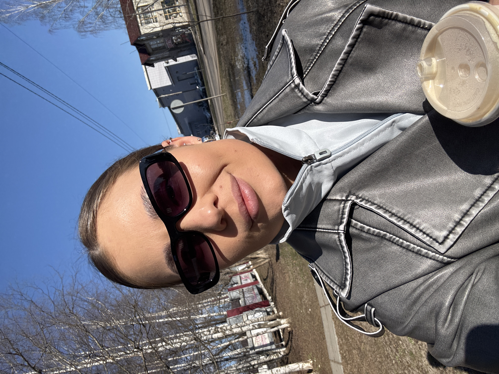 A woman with light brown hair pulled back is wearing sunglasses and a gray leather jacket over a light blue top. She is holding a disposable cup with a lid on it. The background shows a street scene, including a building with a distinctive white circular sign, possibly in Chernushka, Russia, judging by visual elements in the image. Bare trees and patches of ground without snow suggest the onset of spring.