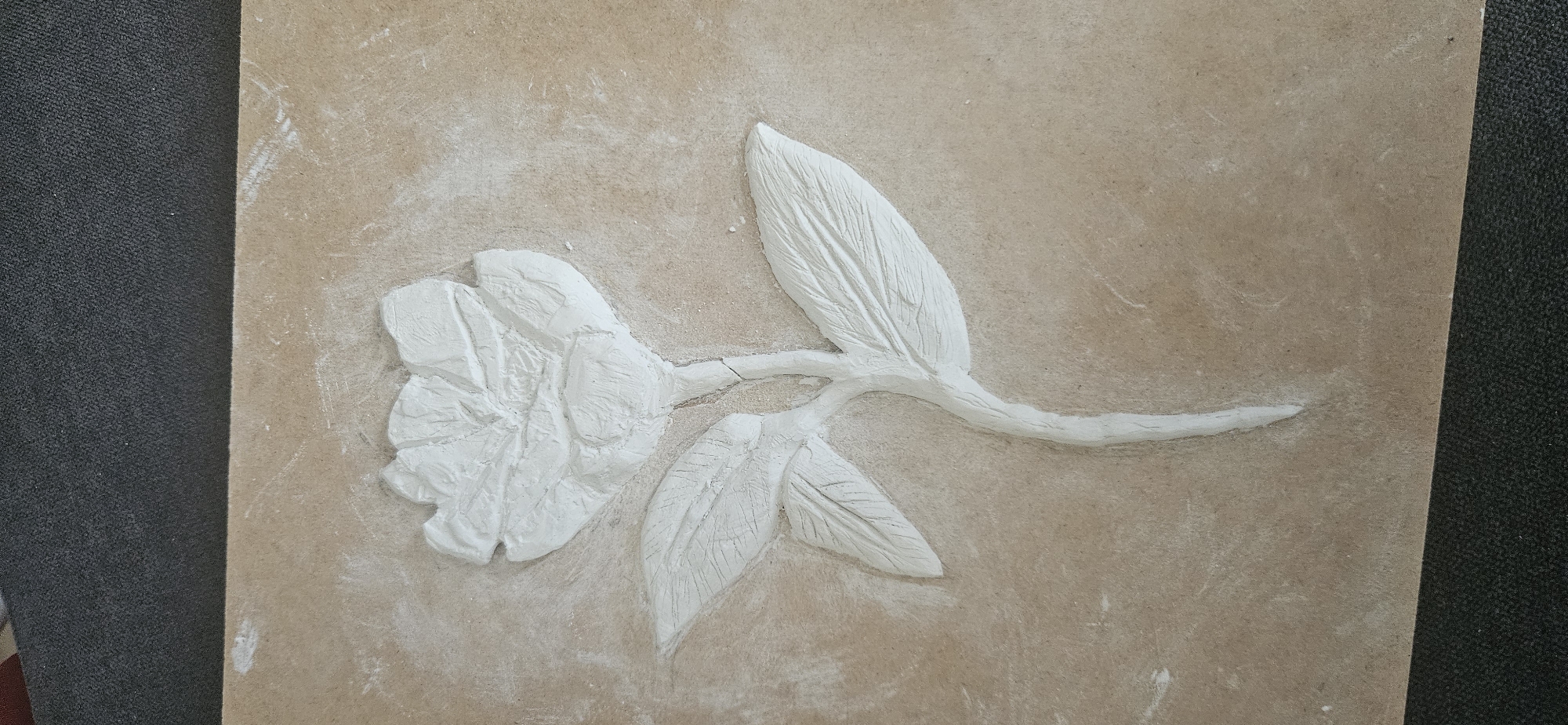The image shows a light brown rectangular slab or board with a sculpted white floral design on it. The floral design features a single bloom with multiple petals, two leaves near the flower, and another leaf extending along a stem. The stem appears to be slightly fractured. The sculpted design is three-dimensional, rising above the surface of the slab. The slab appears dusty, with traces of white residue around the sculpted elements. The environment surrounding the slab is not clearly visible, but a dark, possibly carpeted surface is present on the bottom right edge of the image. There is no indication of the location being Plovdiv, Bulgaria within the image.