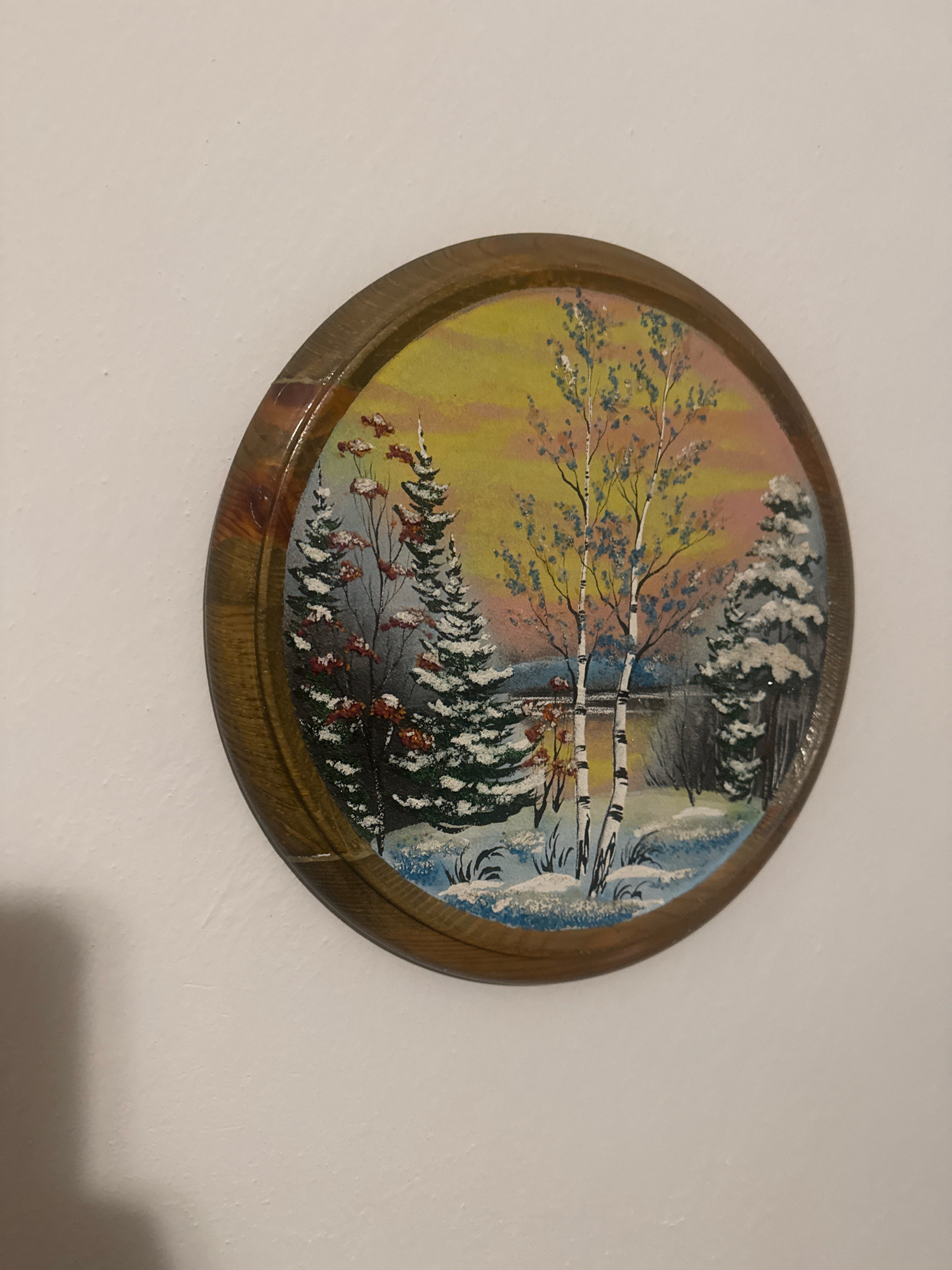 The image shows a circular painting of a winter landscape, framed in wood. The painting depicts snow-covered evergreen trees and birch trees in the foreground, with a calm body of water reflecting the warm hues of a sunset sky. The background includes darker trees and a hint of land. The painting is hung on a textured, off-white wall. The location is not specified in the image itself and therefore cannot be determined, including any specific city in Russia such as Anapa.
