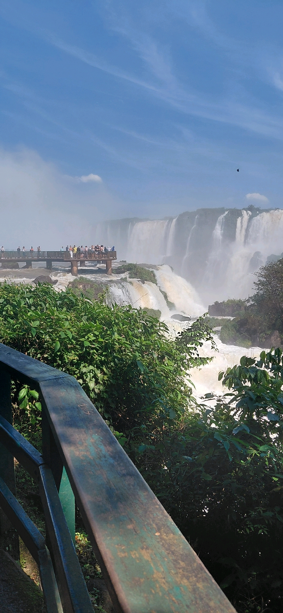 The image shows a panoramic view of the Iguazu Falls, located on the border between Brazil and Argentina in Foz do Iguaçu, Brazil. In the foreground, a weathered wooden railing with a green hue partially obstructs the view. Behind the railing, lush green foliage covers the ground and climbs up towards the falls.
In the middle ground, a wooden boardwalk extends out over the cascading water, with a crowd of tourists gathered on it, observing the spectacle. The boardwalk has a dark brown railing and supports made of concrete pillars. The Iguazu Falls themselves dominate the background, with numerous streams of water plunging down massive cliffs, creating a misty haze that partially obscures the upper sections of the falls. The water appears turbulent and powerful as it descends.
The sky above is a bright blue, with wispy white clouds streaked across it. A small dark bird is visible in flight in the upper right portion of the sky. The overall scene captures the grandeur and natural beauty of the Iguazu Falls, with human presence adding a sense of scale and appreciation to the landscape.