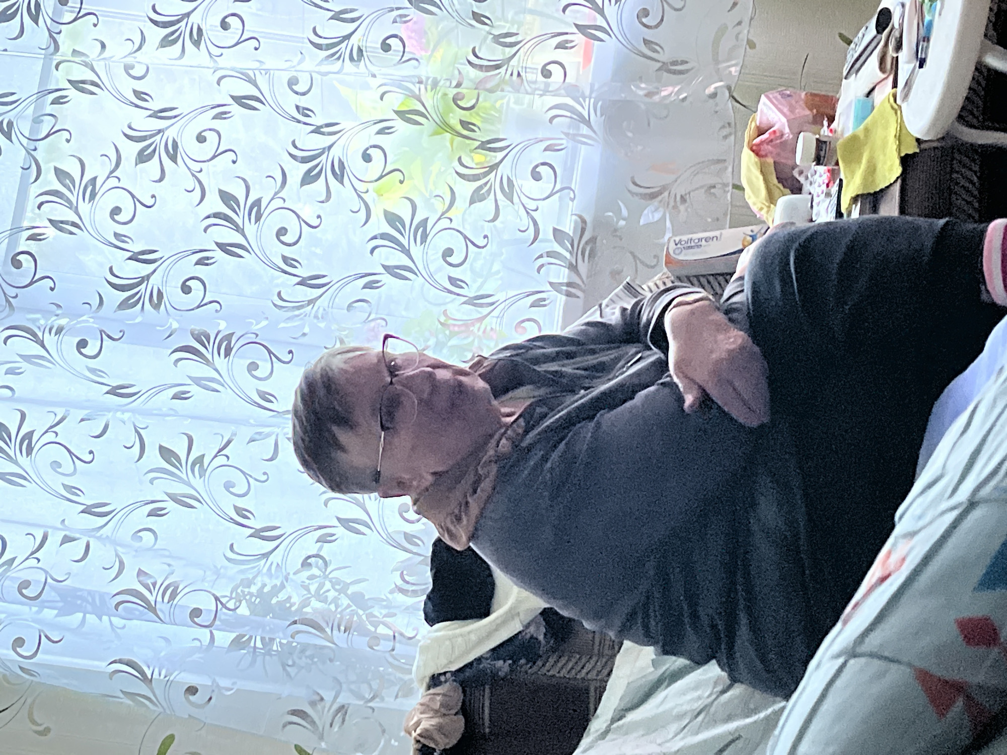 An elderly person with short grey hair and glasses sits on a sofa in front of a window with patterned sheer curtains. The person is wearing a dark grey long-sleeved top and dark trousers. The person's arms are crossed. To the left of the person, on the sofa, are several items of clothing, including a folded white blanket and a dark garment. To the right of the person, on a small table or surface, are various items including a box of medicine, a white bottle, a pink plastic bag, a yellow cloth, and a remote control. The setting appears to be a domestic indoor environment, likely a living room. The overall lighting is soft and diffused, suggesting natural light coming from the window. The image is captured from a medium-wide shot, focusing on the person and their immediate surroundings.