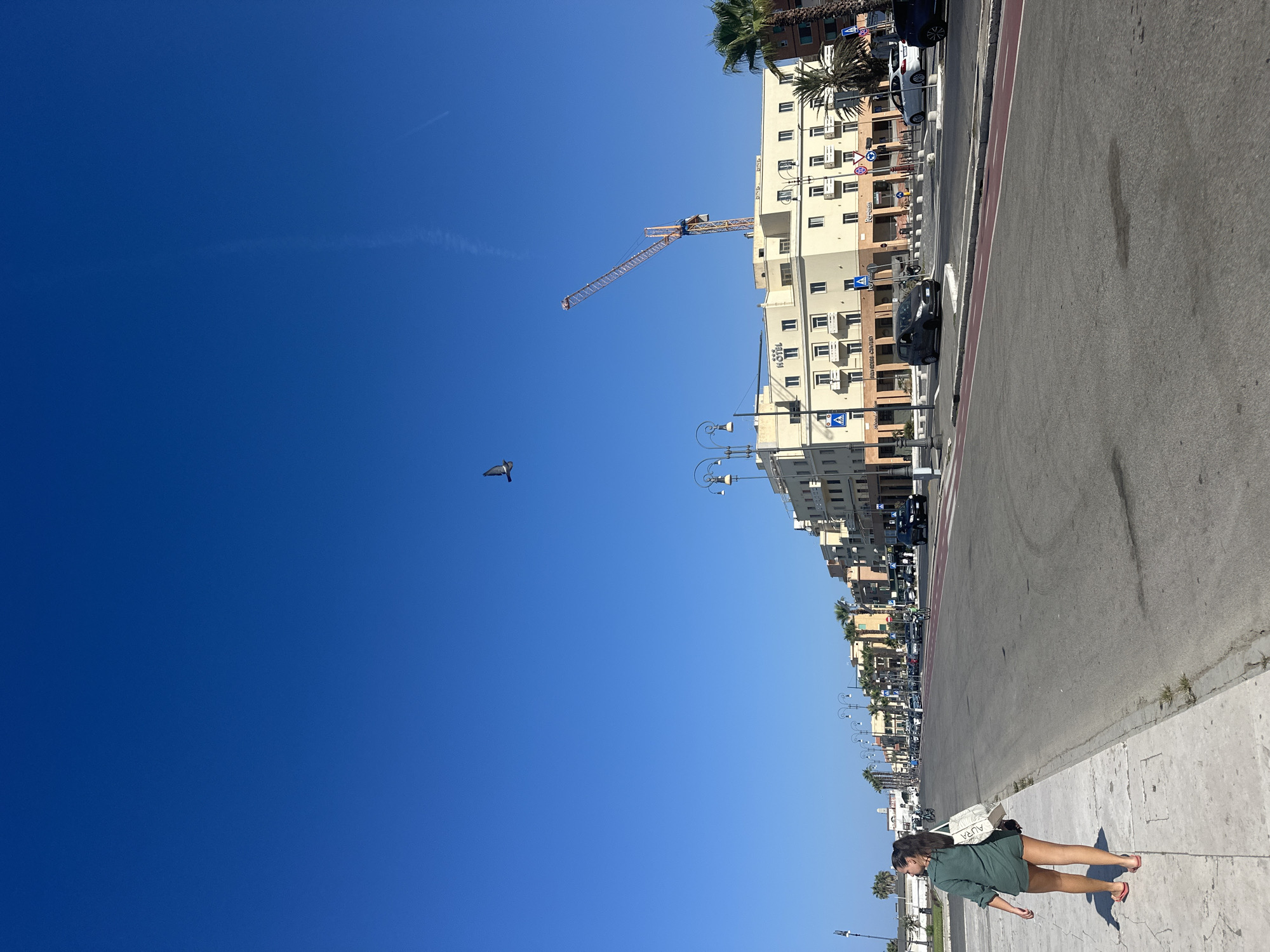 A young woman with dark hair pulled into a ponytail walks on a sidewalk along a street in a coastal city. She is wearing a green shirt and shorts and carrying a white tote bag. The street is lined with buildings, palm trees, and streetlights. A construction crane is visible in the background. The sky is clear and blue. The scene appears to be daytime. The location is not Rome, Italy; it is likely somewhere along the coast.