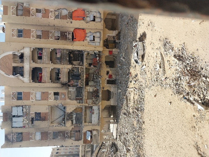 A multi-story residential building in Khan Yunis, Palestinian Territory, displays extensive structural and surface damage. The beige-colored facade is riddled with countless small, circular impact holes across all visible floors. Numerous windows are shattered, missing glass, or have frames blown out, exposing internal apartment spaces, some containing draped fabrics and personal items. Significant charring and localized destruction are evident on multiple lower and middle floors, consistent with fire damage. An orange tarp covers a window opening on the lower right, and a white sheet covers another on a mid-level floor. The ground directly in front of the building is a mix of disturbed sand and dense concentrations of concrete rubble, fractured building materials, and twisted metal debris. A heavily damaged and crushed white sedan is visible amidst the rubble in the central foreground. The scene is illuminated by daylight under a clear sky. No individuals are present in the image.