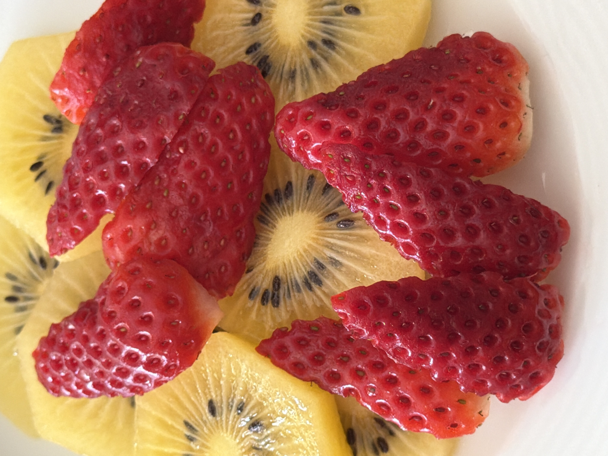 A close-up, overhead shot shows sliced strawberries and yellow kiwi fruit on a white surface. The red strawberry pieces are unevenly cut and scattered among the golden kiwi slices, which are circular and feature a star-shaped pattern of black seeds. The environment is undefined except for the implied white surface, possibly a plate. There are no people visible. The scene focuses solely on the arrangement of the fruit. Esch-sur-Alzette, Luxembourg, is not discernible from the image.