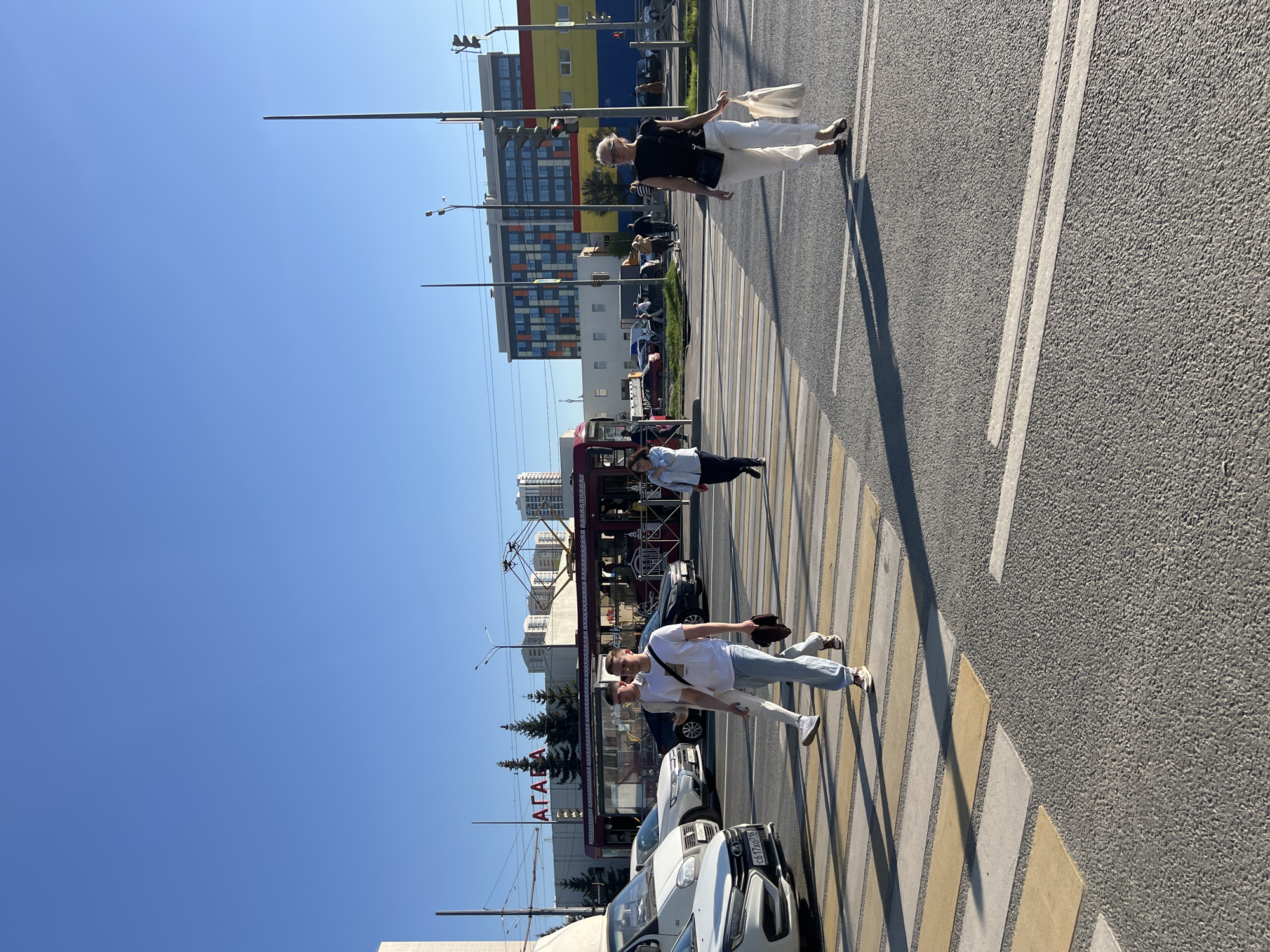 The image shows a city street in Kazan, Russia, with a crosswalk occupied by several pedestrians.
Two women are walking across the crosswalk in the upper portion of the image. One woman is wearing a black shirt and white pants and is carrying a white bag. The other woman appears to be wearing a similar outfit but with a striped shirt.
Further down the crosswalk, a woman in a light-colored shirt and dark pants is lying on the crosswalk.
In the lower portion of the image, two young men in white shirts and light-colored pants are walking. One is carrying a dark-colored hat or bag.
Cars are parked along the street. A red streetcar is present, and the word "АГА" is visible in large red letters on the side of a building. Multi-story buildings and clear blue sky are in the background.