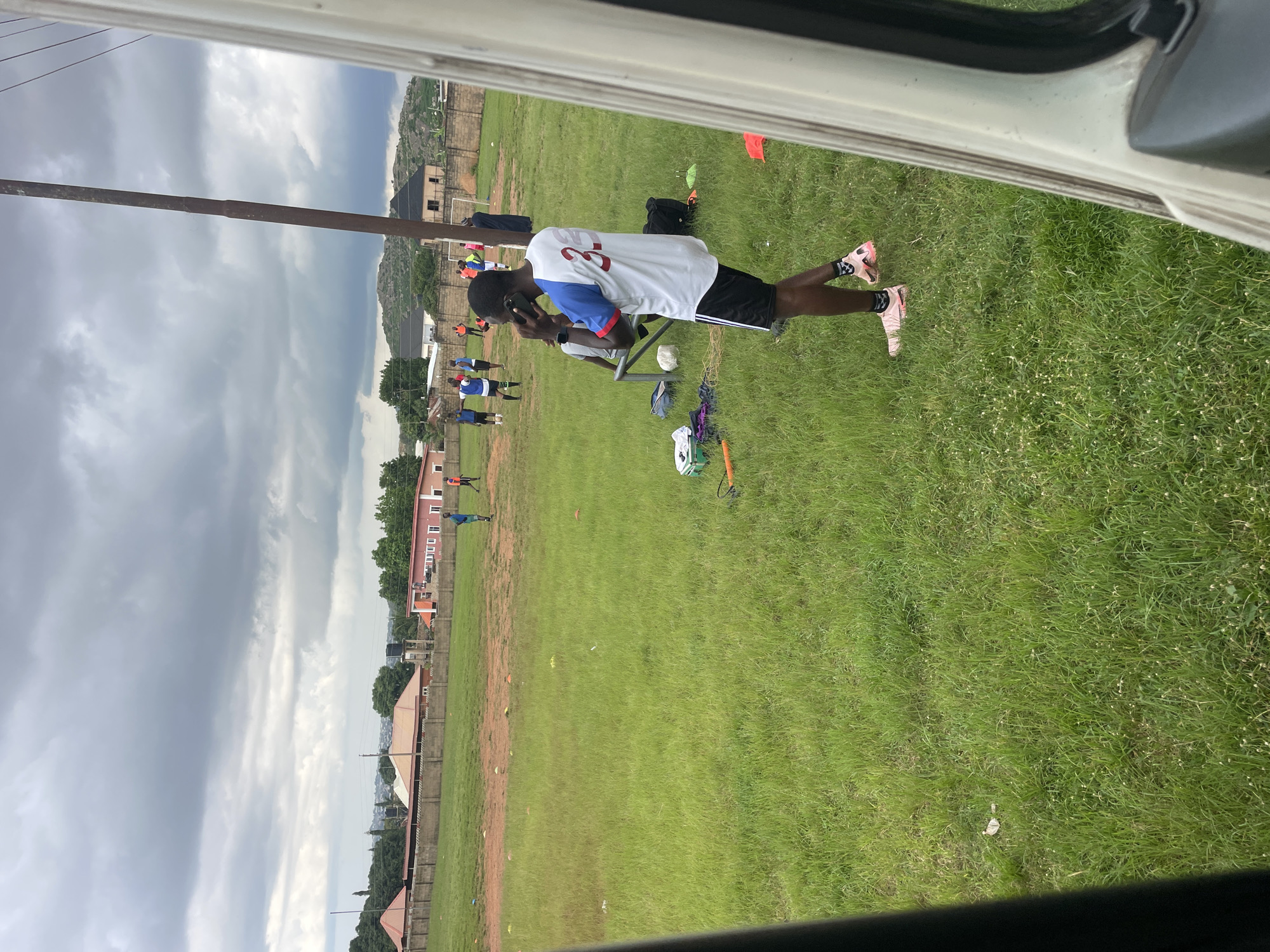 In the city of Jos, Nigeria, a group of young men are gathered on a grassy field, possibly for a sports practice. The sky above is overcast with heavy, dark clouds, suggesting the possibility of rain. The field is well-maintained with lush green grass and scattered orange cones, indicating training exercises. In the foreground, a young man in athletic wear, a white jersey with red numbering "3", and black shorts, is talking on his phone. Several other individuals are scattered across the field, some appearing to be in uniform, suggesting a team or group activity. In the background, residential buildings with red and brown roofs are visible, nestled amongst trees, with a rocky hillside in the distance. The scene is framed by the side of a white vehicle on the right, and a dark, blurry element on the left, suggesting the photo was taken from inside a vehicle.