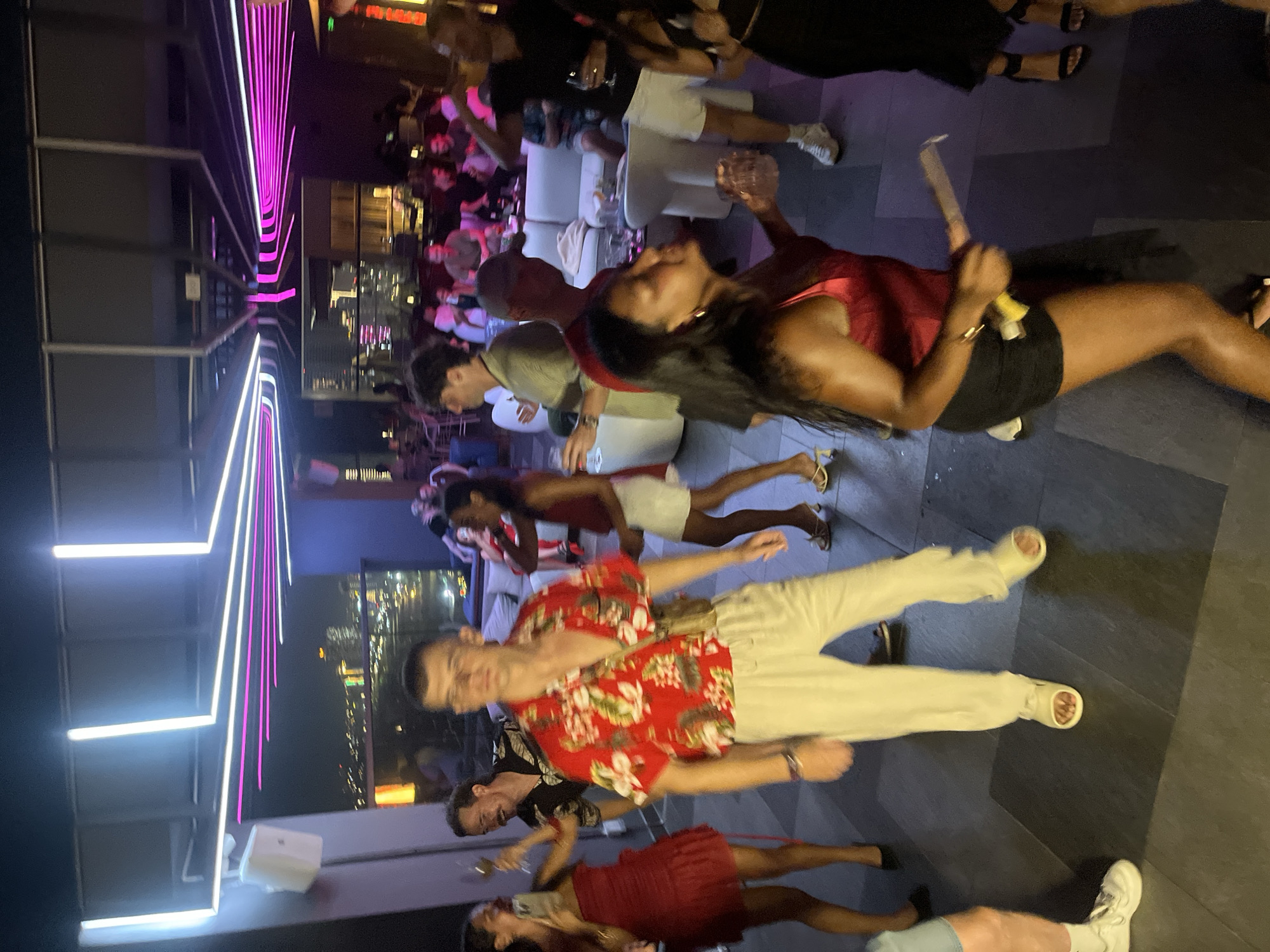 This media file depicts a lively nighttime gathering at a rooftop venue, likely in Đà Nẵng, Vietnam, given the city lights visible in the background. The scene is illuminated by a vibrant display of neon lighting, with prominent pink and white vertical stripes, suggesting a modern and energetic atmosphere.
Several individuals are present, engaged in socializing. In the foreground, a man wearing a red floral shirt and light-colored pants is captured mid-motion, possibly speaking or laughing. Beside him, another man in a dark shirt is also engaged in conversation. Further back, a woman in a red top and black shorts is holding an object, possibly a drink or a prop, and appears to be interacting with someone out of frame. Other people are seen in smaller groups, some seated on white furniture, suggesting a lounge area.
The time of day is clearly night, indicated by the dark sky and the illuminated cityscape. The overall mood seems to be festive and celebratory, with people dressed in casual attire appropriate for a warm evening. The presence of drinks and the social interactions point to an event like a party or a night out.
Notable details include the stylish neon lighting that defines the space, the urban backdrop of Đà Nẵng at night, and the diverse group of people enjoying themselves. The focus on people and their interactions creates a dynamic and engaging visual narrative of a night out in the city.