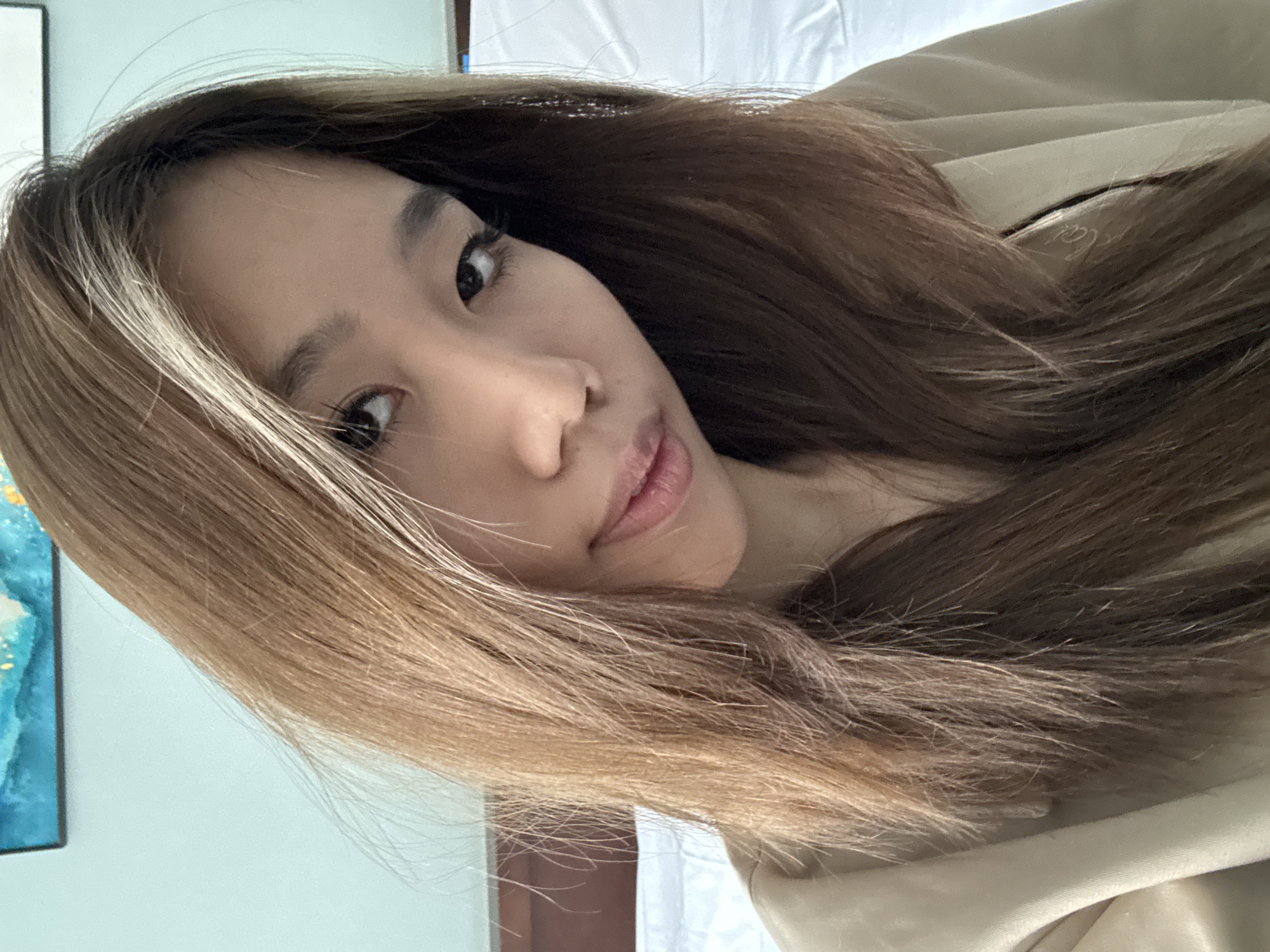 A close-up portrait captures an Asian woman with long, layered brown hair, partially blonde-highlighted near the face, angled slightly towards the camera. Her skin is fair, and her features are delicate. Her eyes are dark, and her lips are full. She wears a neutral-toned top, possibly a jacket or coat. The background is blurred, showing hints of a blue-toned wall on the left and a white sheet or curtain behind her. There is no evidence to suggest the location is Lanzhou, China.