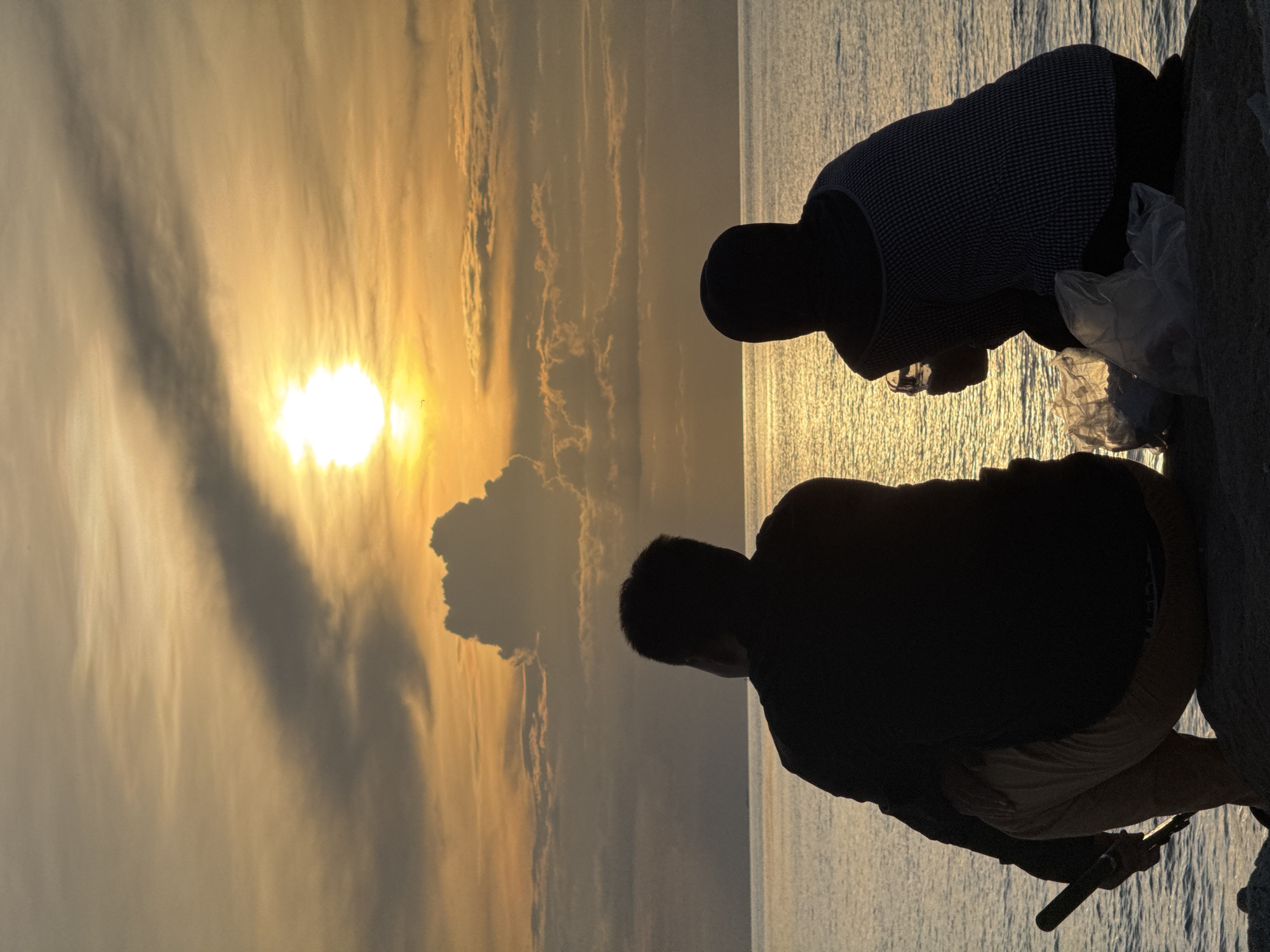 At sunset in Ko Phangan, Thailand, two individuals are silhouetted against the vibrant sky, facing the sea. The person on the left, wearing a dark long-sleeved shirt and light-colored pants, is crouched down, holding a dark, cylindrical object. To their right, the second individual, wearing a patterned top and a headscarf, is also crouched, holding a clear glass. Two plastic bags containing white objects are placed on the ground between them. The sea is calm, reflecting the golden hues of the setting sun, and clouds are scattered across the sky. The foreground consists of rocks and sand.