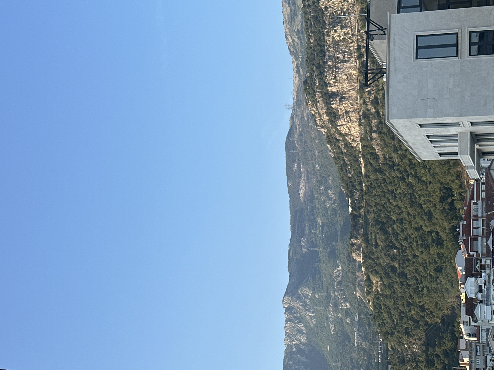 A vertical, eye-level shot captures a serene landscape in Budva, Montenegro, dominated by a clear, pale blue sky that occupies the upper half of the frame. Below, a range of rugged, tree-covered mountains stretches across the horizon, their slopes dotted with intermittent rock formations and a winding road. Faintly visible on the mountain peaks are power lines. In the foreground, a modern, multi-story building with a light gray facade and dark-paned windows stands prominently. To the left and below this building, a cluster of residential buildings with red-tiled roofs and white and beige walls are visible. The overall scene conveys a tranquil, daytime atmosphere with no people or active events depicted.