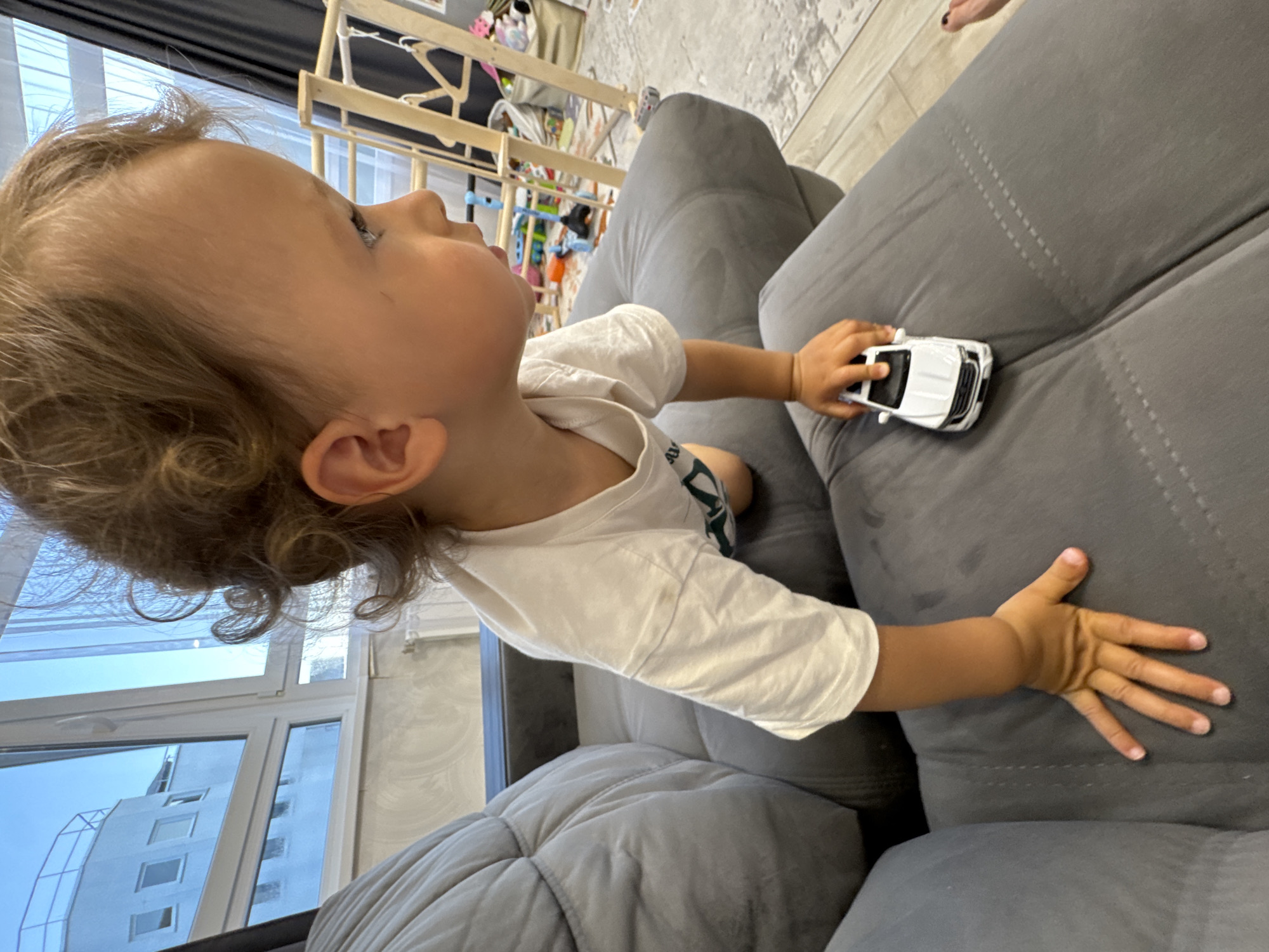 An overhead-angle interior photograph taken in Sochi, Russia, depicts a young child (toddler) crawling across a large, grey upholstered sofa. The child, with light brown curly hair, is seen in profile from the left, wearing a white short-sleeved t-shirt, and holds a white toy SUV in their right hand. Their left hand is placed on the sofa cushion for support. The child is looking towards the right side of the frame, beyond the visible area.
In the background, a large window reveals a white multi-story building with multiple rectangular windows under a light blue sky. Below the window, a white radiator is visible, and to the right, dark grey curtains are drawn open. The interior features light-colored walls with a subtle texture and a light-toned floor covered by a patterned rug. Further in the background, a child's play area is visible, including a wooden climbing structure, various colorful toys (e.g., a blue toy, scattered items), and a fabric storage basket filled with objects. A partial view of an adult's foot with dark painted toenails is visible on the far right edge of the sofa.