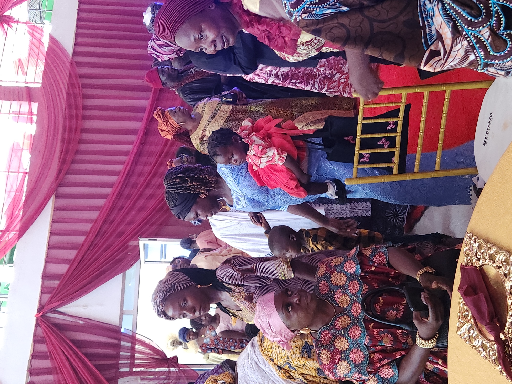 The photograph captures an indoor event in Barden Yana, Nigeria, characterized by vibrant decorations and a gathering of people. The scene is adorned with rich, dark red drapery, which hangs from the ceiling and walls, creating a festive and somewhat formal atmosphere. The lighting appears to be a mix of ambient and possibly artificial light, contributing to the bright, slightly overexposed quality of the image.
In the foreground, a woman is seated at a table, holding a mobile phone and a black handbag. She is wearing a colorful dress adorned with floral patterns and jewelry, including bracelets and a necklace. To her left, another woman, also richly dressed, is looking to the side. A small child, dressed in a bright red outfit, is being held by a woman in a blue dress. The child is looking away from the camera.
Further into the scene, a group of individuals are standing and conversing. One man is wearing a traditional dark outfit and a red cap. Another woman is dressed in an orange headwrap and a patterned yellow and brown dress. The background shows more people, some of whom appear to be children, and additional draped fabric. The overall impression is of a celebratory occasion, possibly a wedding or a similar social gathering, taking place within a decorated indoor venue. The presence of traditional attire suggests a cultural context specific to Nigeria.