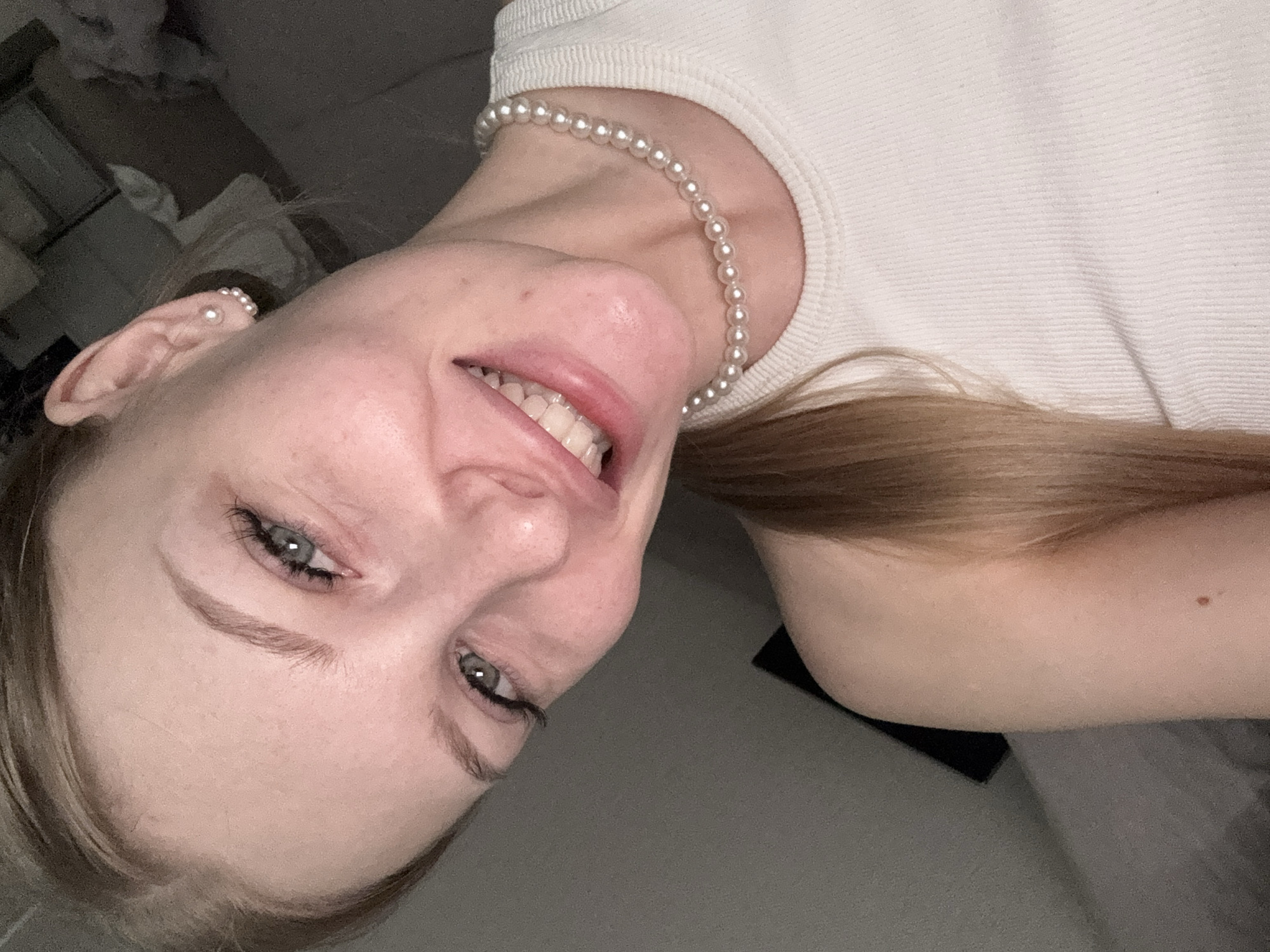 A close-up, tilted, selfie-style photograph depicts a fair-skinned female with light brown hair. She is smiling and wearing a pearl necklace and pearl earrings. Her hair is pulled back on one side. The background is blurred and indistinct, showing glimpses of what appears to be bedding and dark furniture. There is no indication of the location being Perm, Russia.