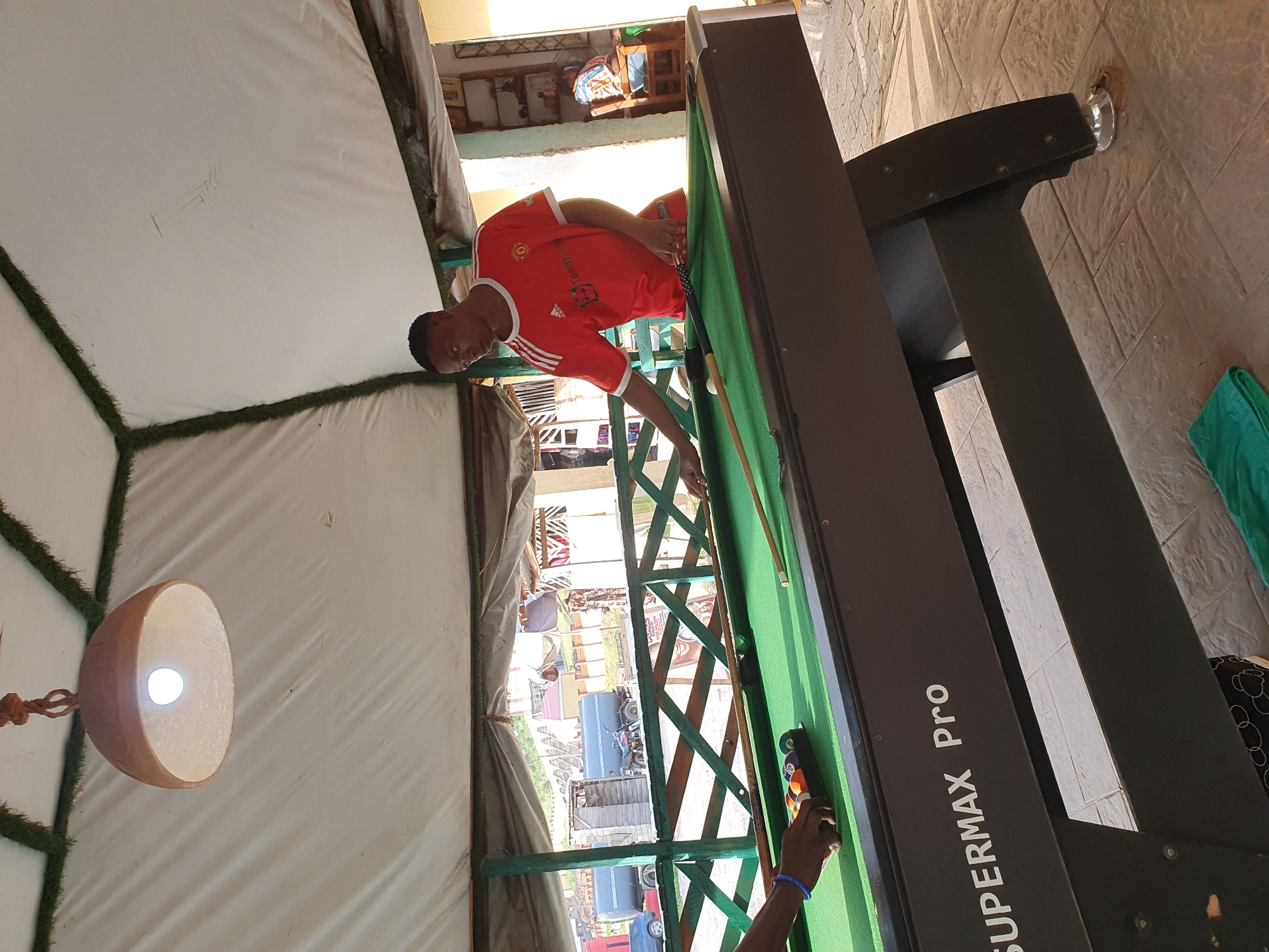 A person is playing pool on a "SUPERMAX Pro" table. They are wearing a red t-shirt with white stripes on the sleeves and orange shorts. The pool table is green with several balls arranged. A pool cue is in the person's hand, suggesting they are about to take a shot. The setting appears to be an outdoor area, possibly a patio or covered space, with a tiled floor and a white tent-like structure in the background. A hanging lamp with a light bulb is also visible in the frame. The image is taken outdoors during the day, indicated by the bright lighting. The overall mood seems relaxed, with the person focused on the game.