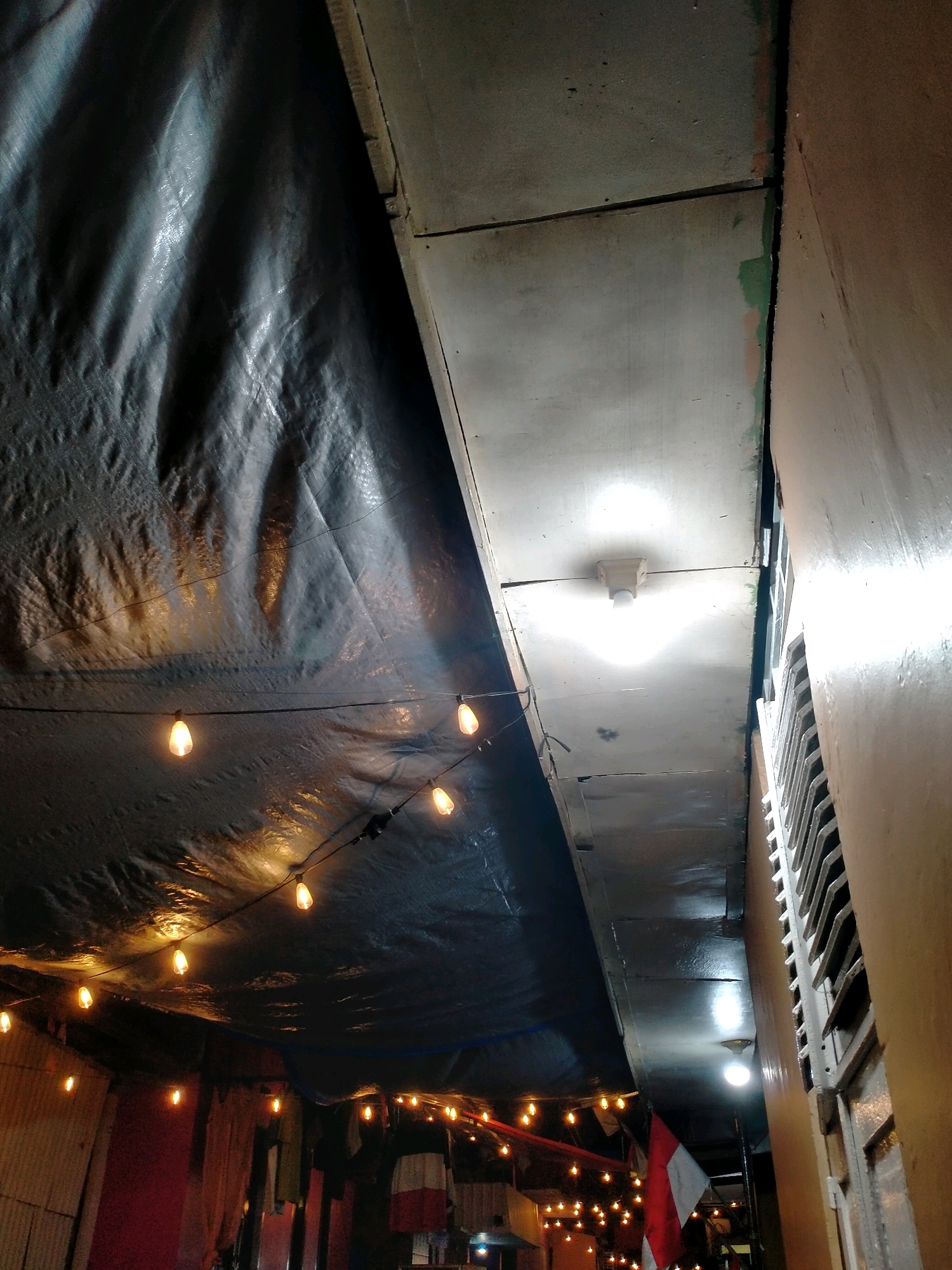The image depicts an outdoor scene at night, likely in a city, given the presence of artificial lighting and the urban-style construction visible. The primary focus is on a makeshift roof made of dark, crinkled tarp, draped over a wooden structure. Strings of warm-toned, exposed bulbs are strung across the tarp, providing ambient illumination and creating reflections on its surface.
To the right of the tarp, a light-colored, possibly metal, ceiling or overhead structure is visible. This structure appears to be made of panels and has a single, bright light fixture attached to it, casting a concentrated glow. A narrow gap is visible between this structure and an adjacent wall, which is painted a light brown or tan color and has a visible ventilation grate.
In the background, below the tarp, the scene opens up to reveal more of the environment. Several other strings of lights are present, suggesting an area that is set up for events or an outdoor marketplace. Red and white Indonesian flags are partially visible, hanging down. Clothes or fabrics are also seen hanging, possibly for sale or display. The overall impression is one of a dimly lit, improvised outdoor space.
While the image shows elements suggestive of a city, such as building structures and lighting, specific details to pinpoint the exact location within Ambon, Indonesia, are not explicitly present. The scene appears to be an outdoor market or gathering space, possibly set up for an evening event. There are no people visible in the image.