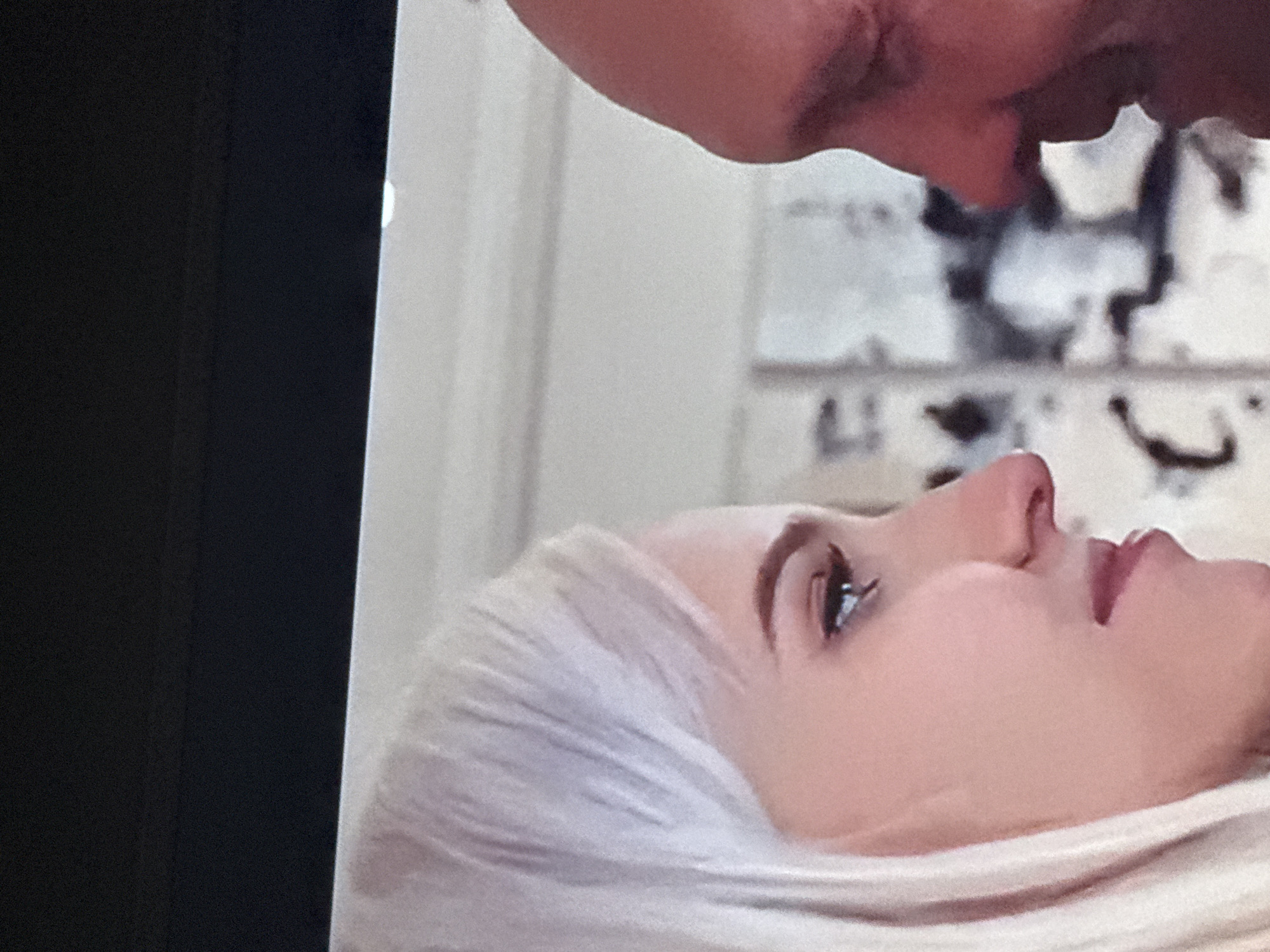 The image shows a close-up shot of two people. The lower person is a woman with fair skin and blonde hair. She is lying on her back, with her face visible in profile. The upper person's face is cropped at the top of the image, showing only part of their nose and upper lip. They appear to be leaning towards the woman. The background is blurry, but it seems to be an indoor setting, possibly a room with wallpaper or a patterned wall. The image has been taken from a screen, as indicated by the black border on the left. There is no evidence or indication that this scene has any relation to the city of Mytishchi, Russia.