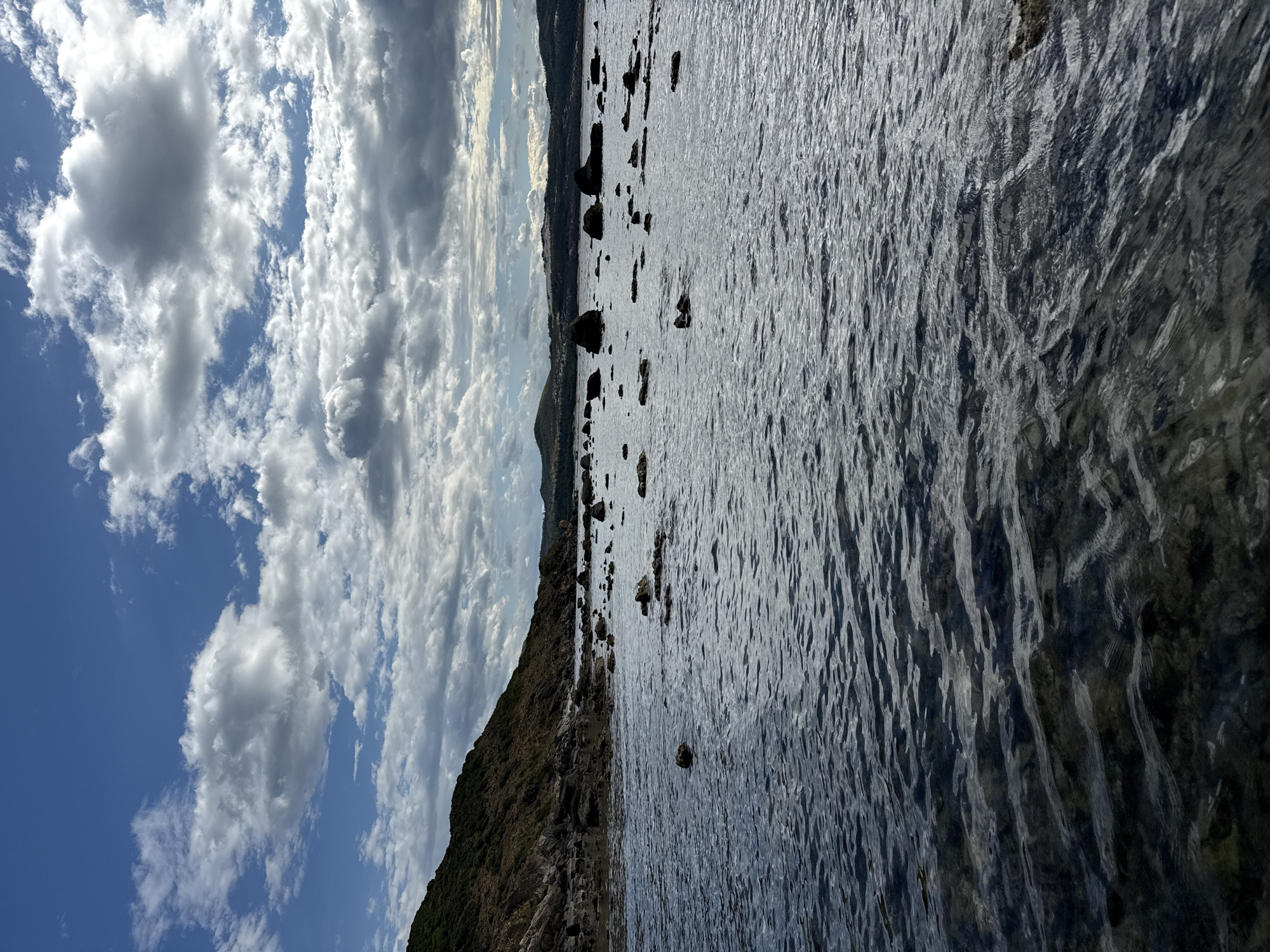 The image shows a body of water with numerous rocks scattered across its surface and along the shoreline. The water is dark and appears to be slightly rippled, with sunlight reflecting off its surface in places. In the background, there is a gently sloping, vegetated hill on the left and rolling hills in the distance on the right. The sky is partly cloudy with dramatic, textured clouds against a bright blue sky. The scene appears to be a coastal or lakeside environment. The location is identified as Từ Nham, Vietnam. There are no people or visible structures in the image.
