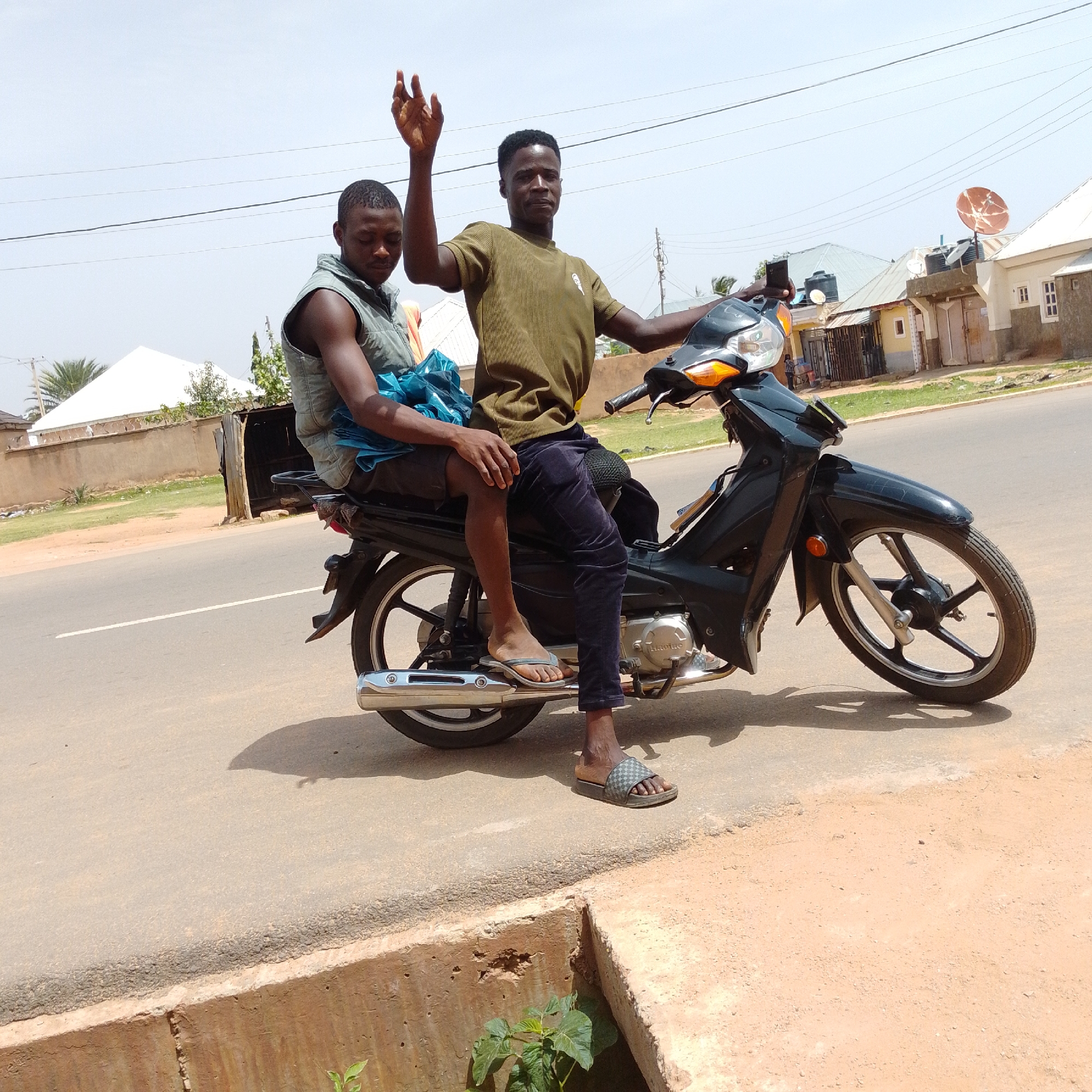 Two young men are seated on a black motorcycle on a paved road in Gombe, Nigeria. The man in the front is wearing a green, ribbed, short-sleeved shirt and dark trousers, and has his right arm raised with his hand open, palm facing forward, as if waving. He is looking towards the camera. The man behind him is wearing a light grey sleeveless vest over a blue garment, and dark shorts. He is looking down and to his left. The motorcycle has a black frame, orange turn signals, a silver exhaust pipe, and a black seat. The road is asphalt with a white dividing line visible. To the right of the road is a dirt shoulder. In the background, there are several buildings with light-colored roofs, some with water tanks on top. One building has a satellite dish on its roof. There are power lines visible in the sky. The overall environment appears to be a street in a residential or commercial area.