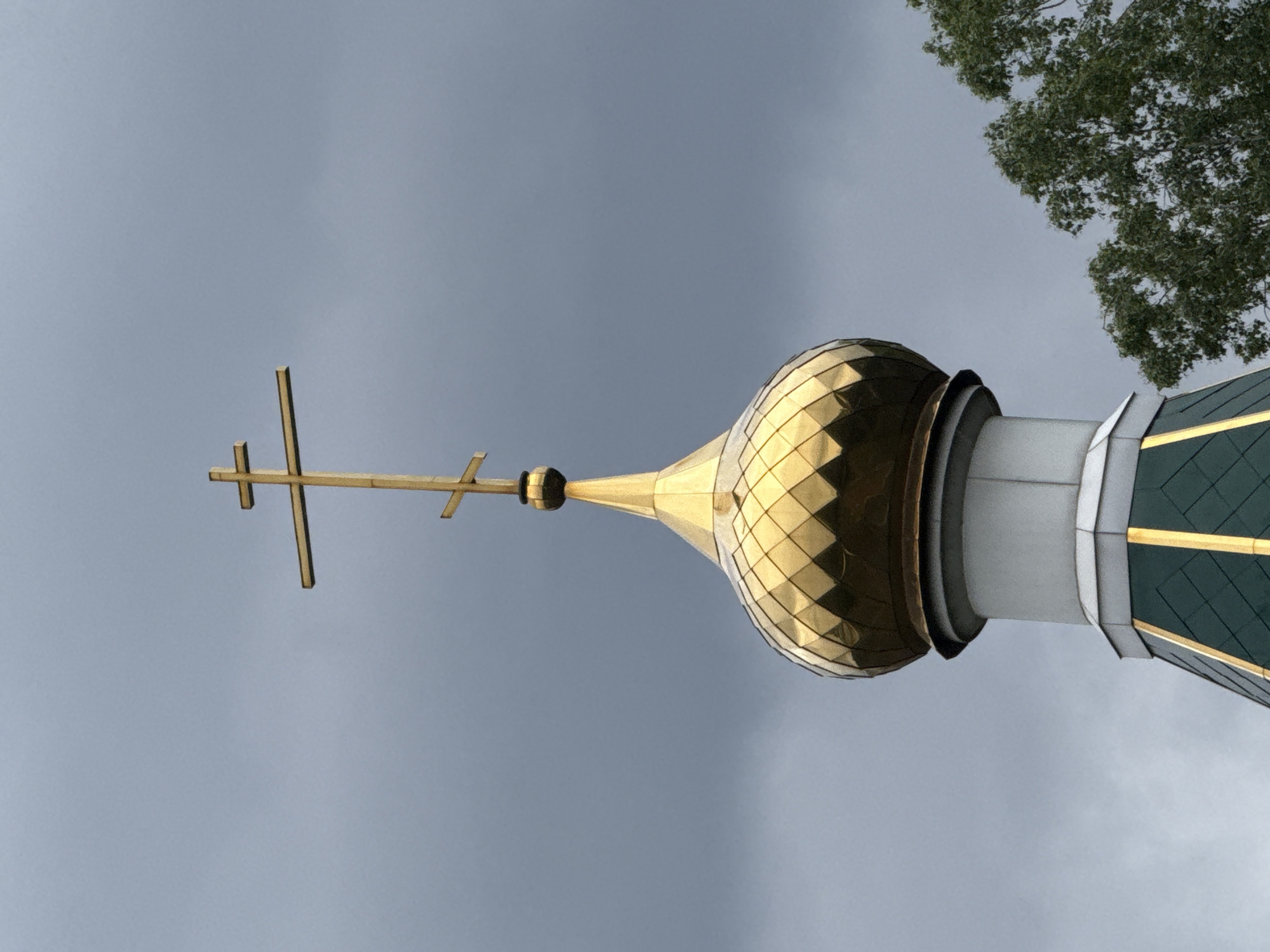 The image displays the upper section of an Eastern Orthodox church, photographed from a low angle against an overcast sky in Chaikovsky, Russia. The primary subject is a shimmering golden onion dome, featuring a mosaic-like pattern of diamond-shaped tiles that catch and reflect light. Ascending from the dome is a slender, golden spire, which is crowned by a three-barred Orthodox cross, also in gold.
Below the distinctive golden dome, the church structure includes a cylindrical, light grey or white section, transitioning further down to a green roof area. This green roofing also appears to have subtle patterned details, possibly with thin gold lines or trim. The sky in the background is a uniform, dull grey, suggesting a cloudy or overcast day. In the upper right corner, a segment of a tree with green foliage is visible, indicating an outdoor setting. No people are present, and no specific activities are taking place. There is no visible text in the image. The architectural style, particularly the onion dome and Orthodox cross, are characteristic of churches found in Russia.