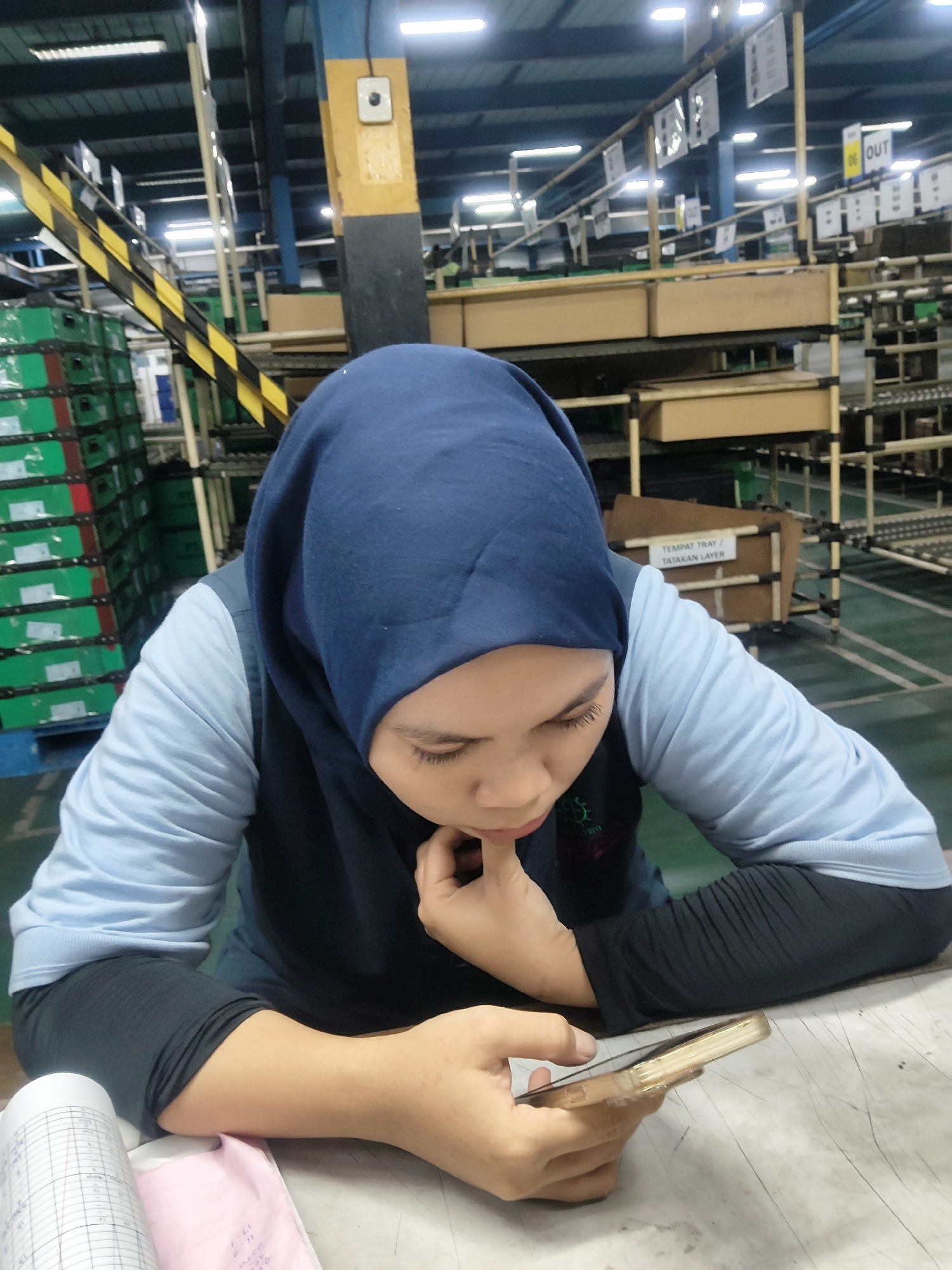 A young woman wearing a dark blue hijab and a light blue long-sleeved shirt is looking down at her mobile phone while resting her chin on her hand. She is seated at a grey table in what appears to be a factory or warehouse environment. In the background, there are stacks of green boxes, shelves with brown cardboard boxes, and overhead fluorescent lighting. A yellow and black striped conveyor belt is also visible. The general atmosphere suggests an industrial setting. The text on a sign in the background reads "TEMPAT TRAY / TATAKAN LAYER", which translates to "TRAY PLACE / LAYER BASE". The location is identified as Mekarmukti, Indonesia, likely within an industrial zone or factory.